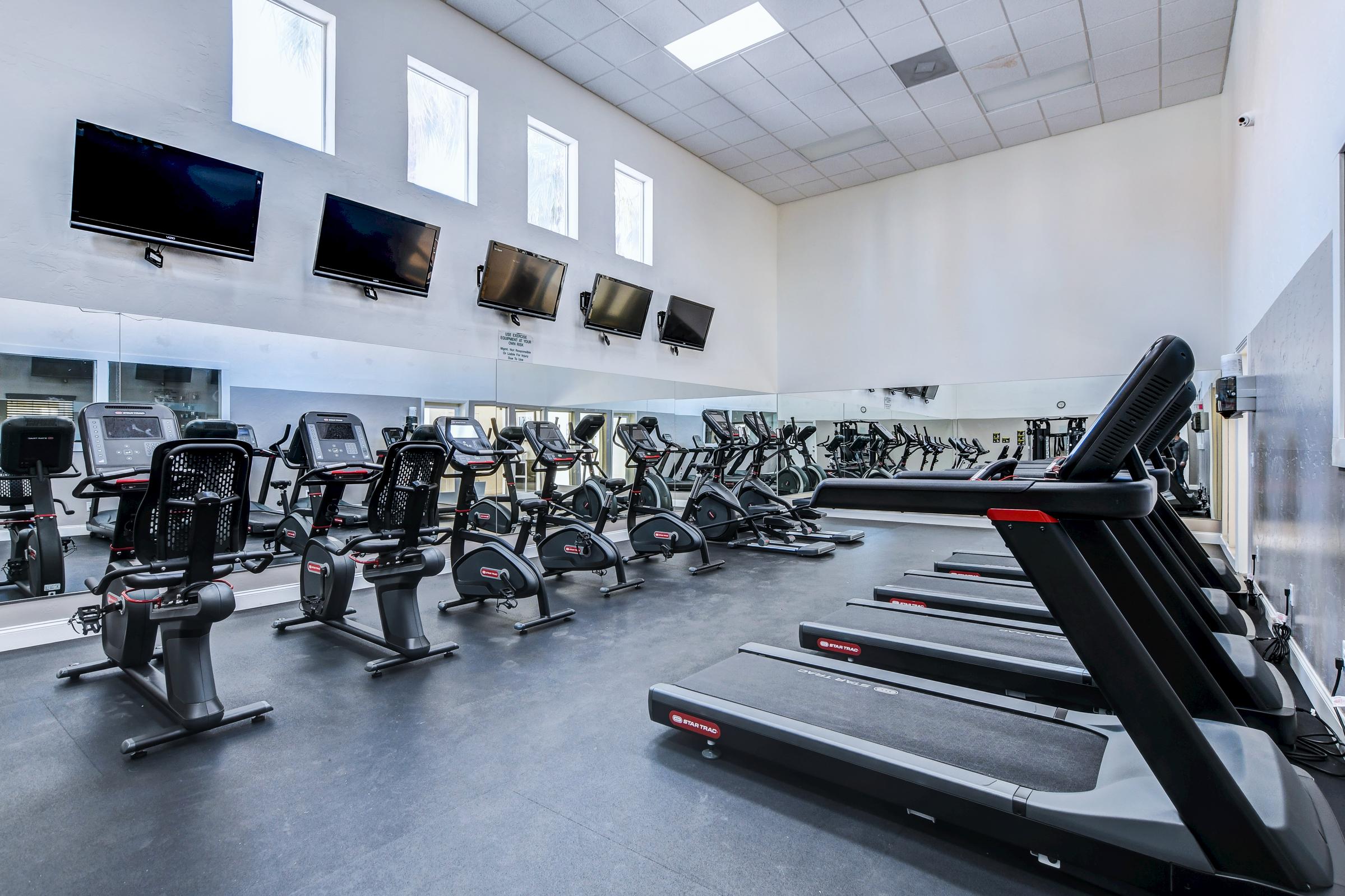 A modern gym interior featuring multiple treadmills and exercise bikes. The space has large mirrors and several televisions mounted on the wall. Bright windows allow natural light to enter, creating an inviting atmosphere for workouts. The gym equipment is well-organized and maintained.