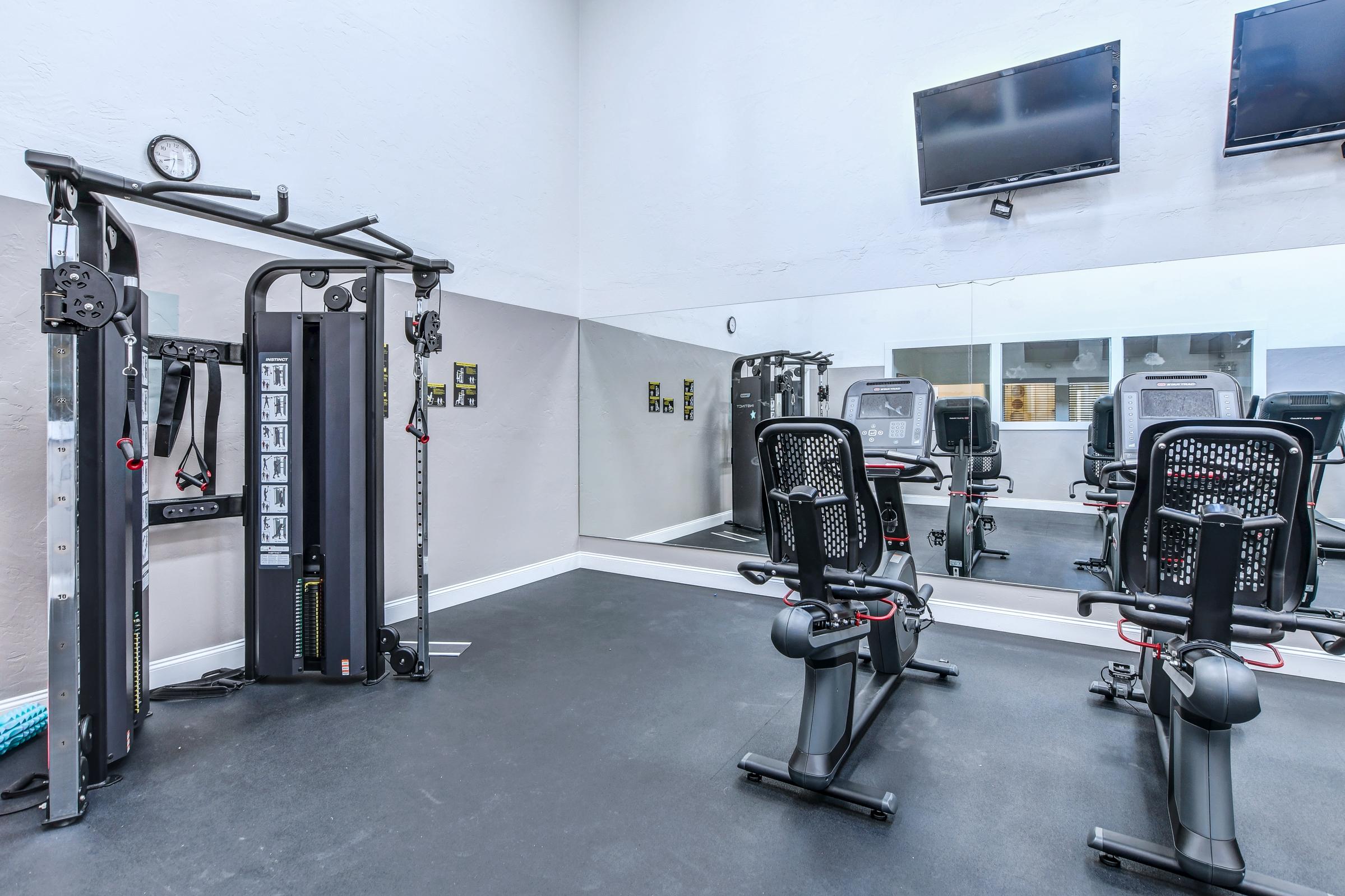 A modern gym interior featuring a cable machine on the left, two exercise bikes in the foreground, and two wall-mounted TVs. Mirrors line one wall, reflecting the equipment, creating an open and bright training space. The flooring is dark and equipped for fitness activities.