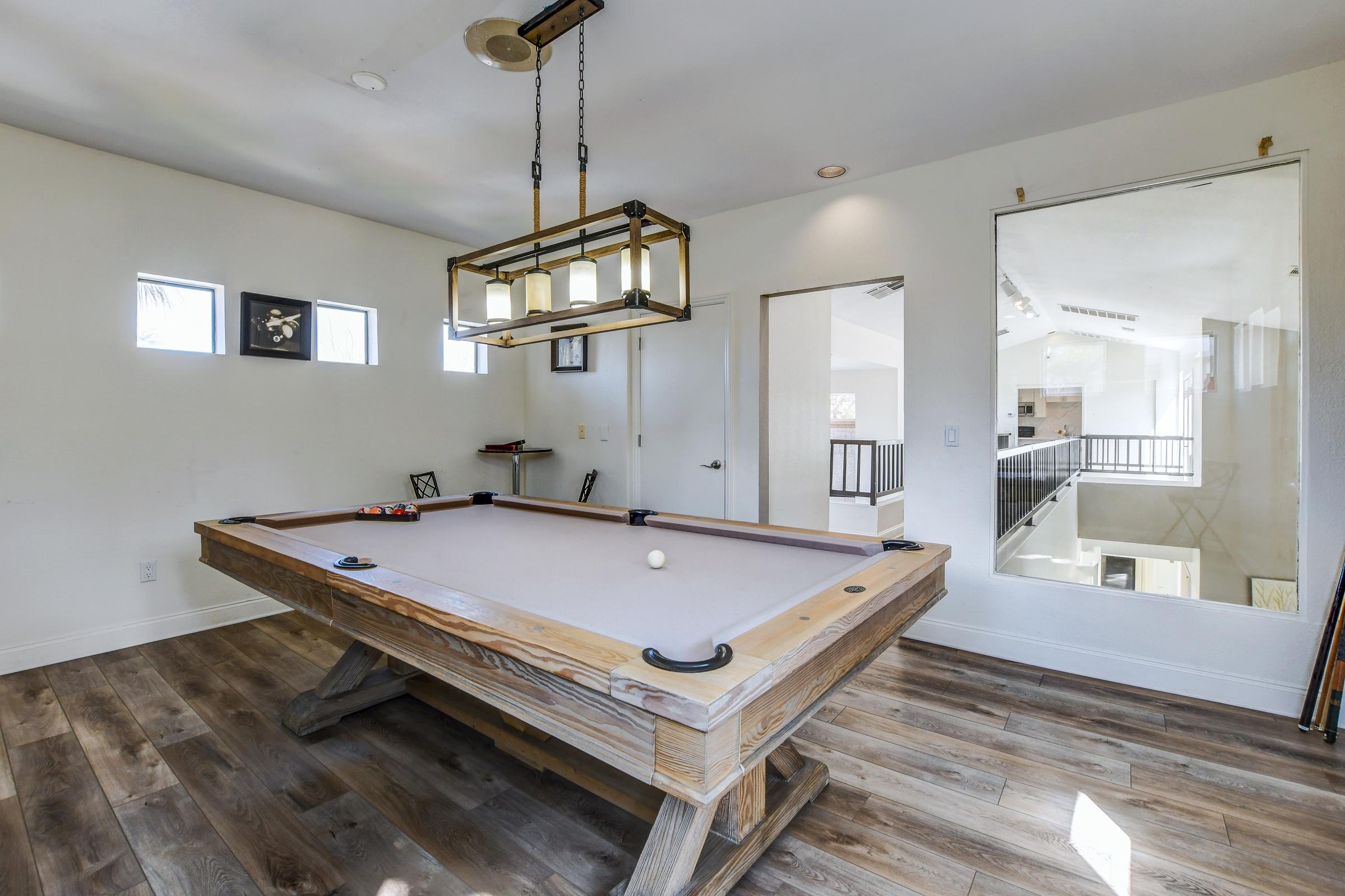 A bright and spacious room featuring a wooden pool table with a white felt surface. The walls are painted white, and there are large windows allowing natural light. A light fixture hangs above the table, and artwork is displayed on one wall. There is a doorway leading to another part of the house visible in the background.