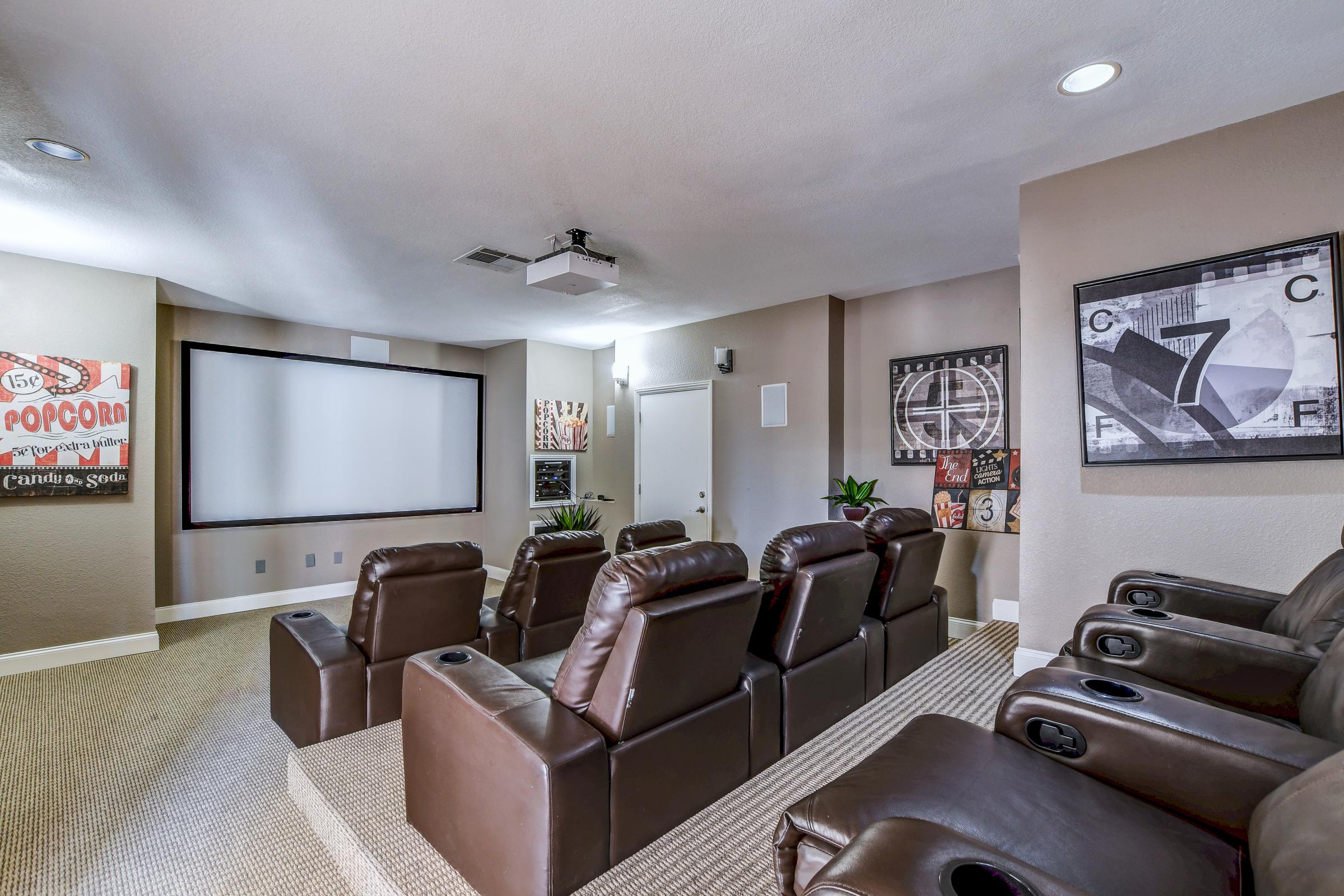 A cozy home theater featuring plush brown recliners arranged in front of a large screen, with a projector mounted on the ceiling. The walls are adorned with framed movie-related artwork and a subtle carpeted floor. 