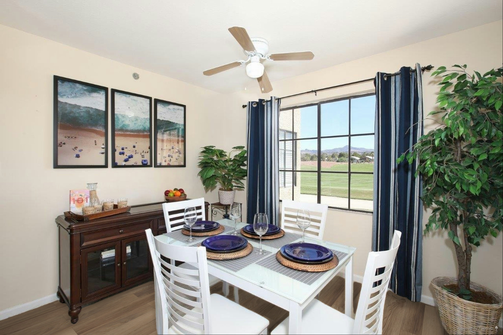 A bright dining area featuring a white table set for four with blue plates and glasses. There is a fan overhead and large windows with blue curtains offering a view of greenery outside. A sideboard displays a fruit bowl and decor. Wall art depicts a beach scene in three panels.