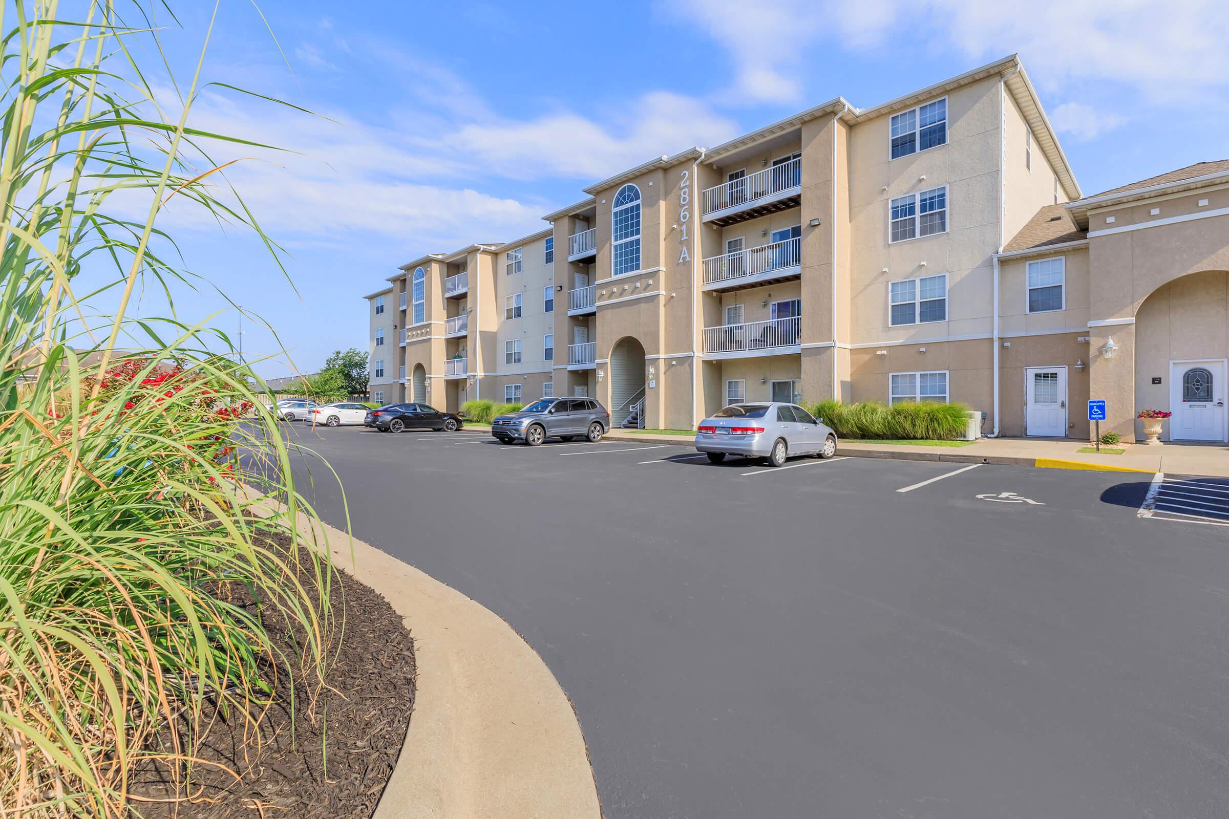 A modern apartment complex with multiple stories, featuring balconies and large windows. The foreground shows a well-maintained parking lot with several cars parked. Lush green plants and landscaping line the edge of the image, contributing to a welcoming atmosphere under a clear blue sky.