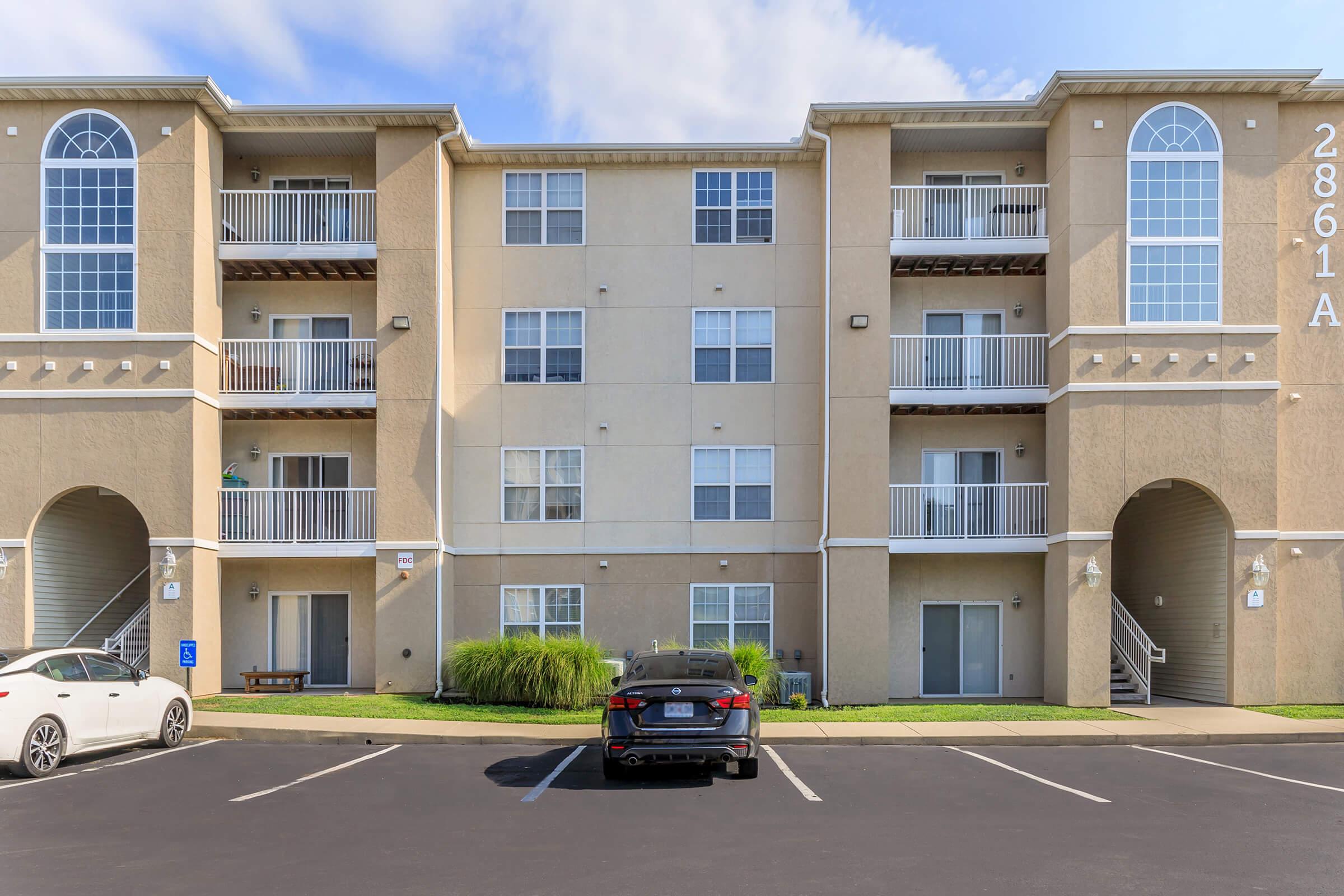 A three-story apartment building with multiple balconies, featuring beige walls and large windows. In the foreground, there are parking spaces with a black car and a white car. Lush grass and small shrubs are visible near the building, under a partly cloudy blue sky.