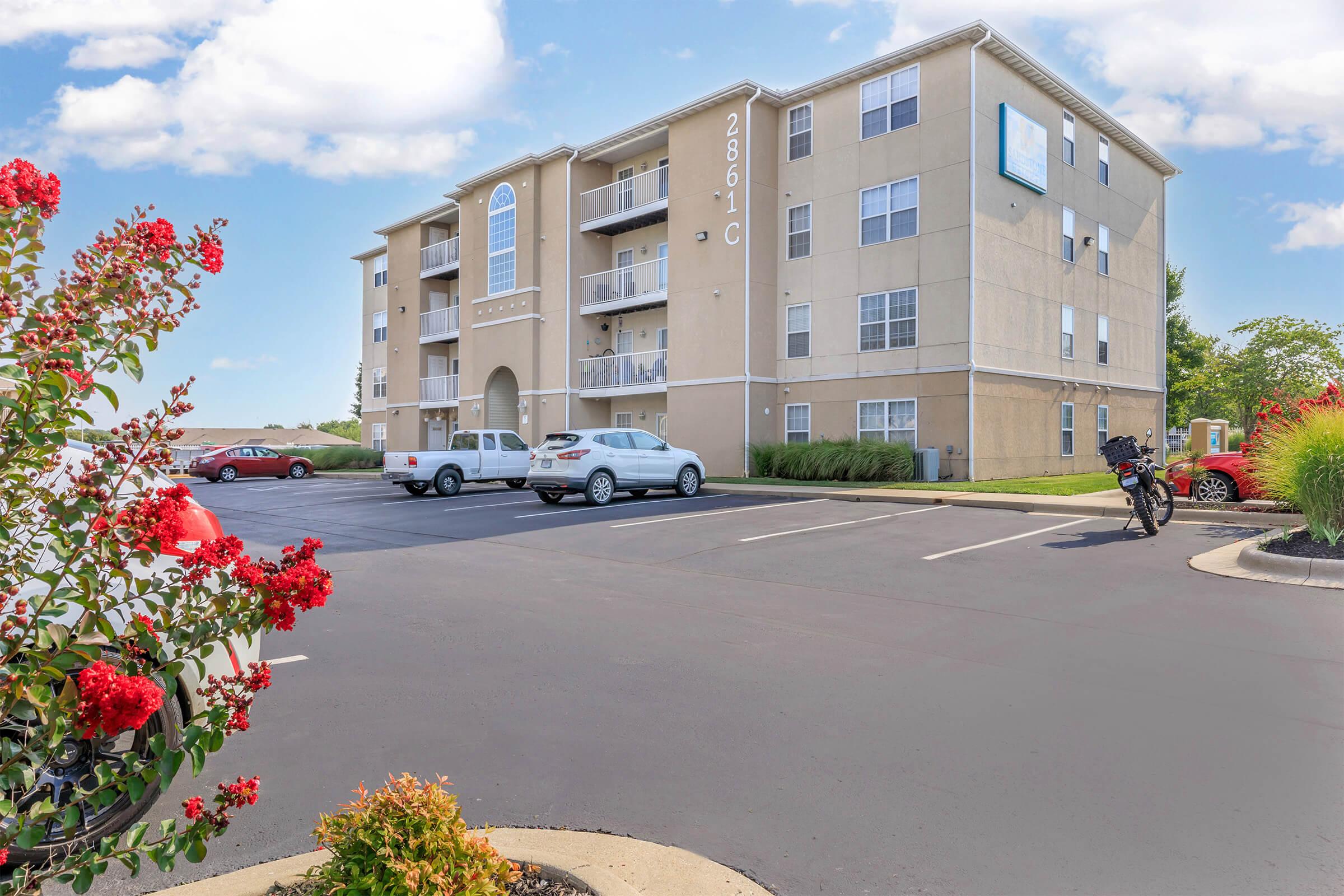 A three-story apartment building with beige exterior and multiple balconies. In the foreground, red flowers are visible along the edge of a paved parking lot, which has several parked vehicles. A motorcycle is parked nearby, and there are green shrubs in the area, set against a clear blue sky with fluffy white clouds.