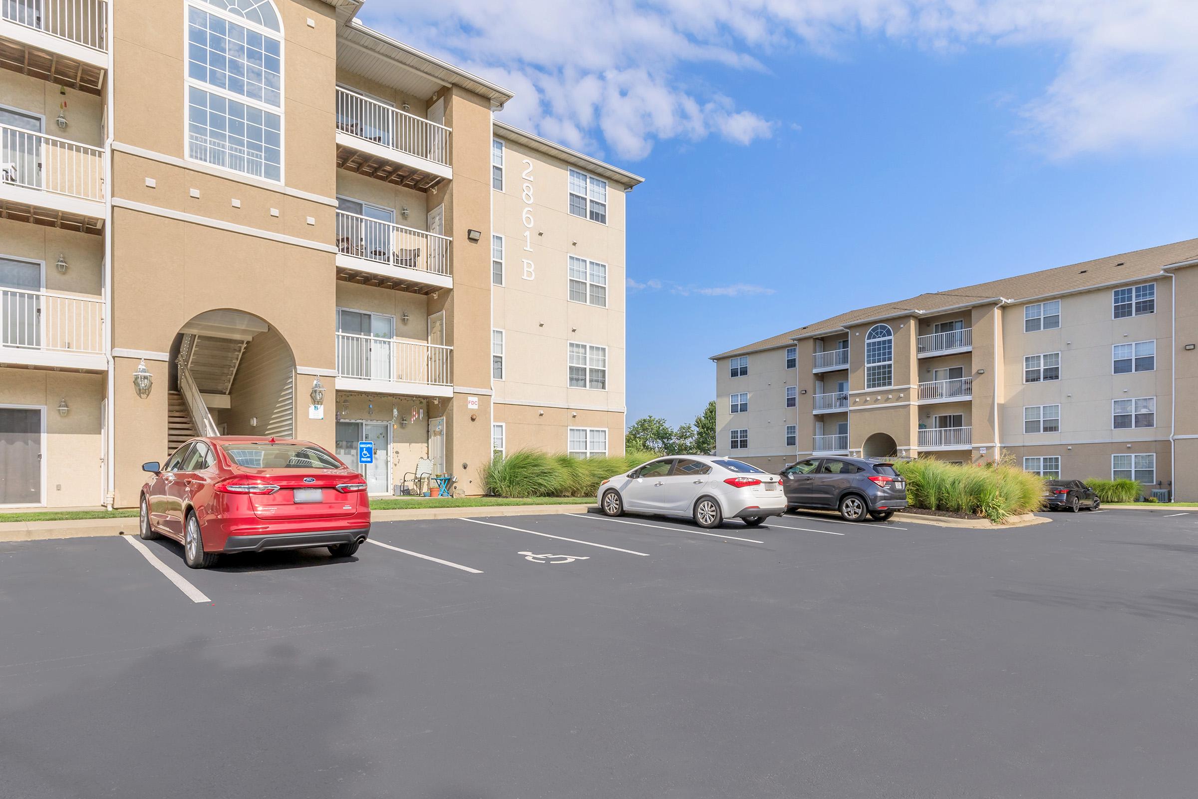 A view of a multi-story apartment building with balconies, showcasing a parking area with several cars. The sky is bright with scattered clouds, and there are grassy areas surrounding the parking lot. The building features large windows and a prominent entrance.