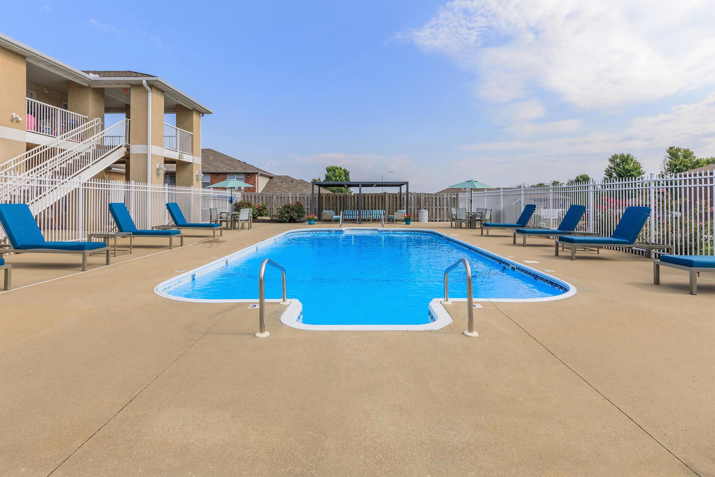 A clear blue swimming pool surrounded by lounge chairs on a sunny day. The pool area features a concrete deck, white fencing, and a gazebo in the background. The scene is set in a residential complex, with buildings visible on one side and trees in the distance.