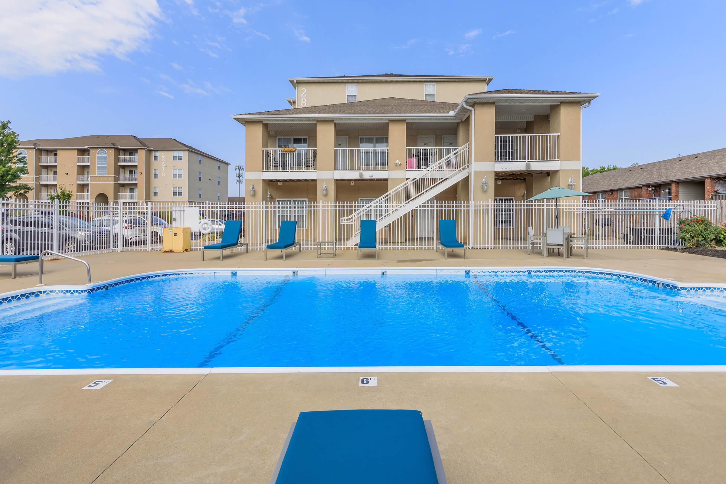 A swimming pool with clear blue water is situated in front of a multi-story building. The pool area features lounge chairs and a surrounding fence. In the background, additional residential buildings are visible under a clear blue sky.