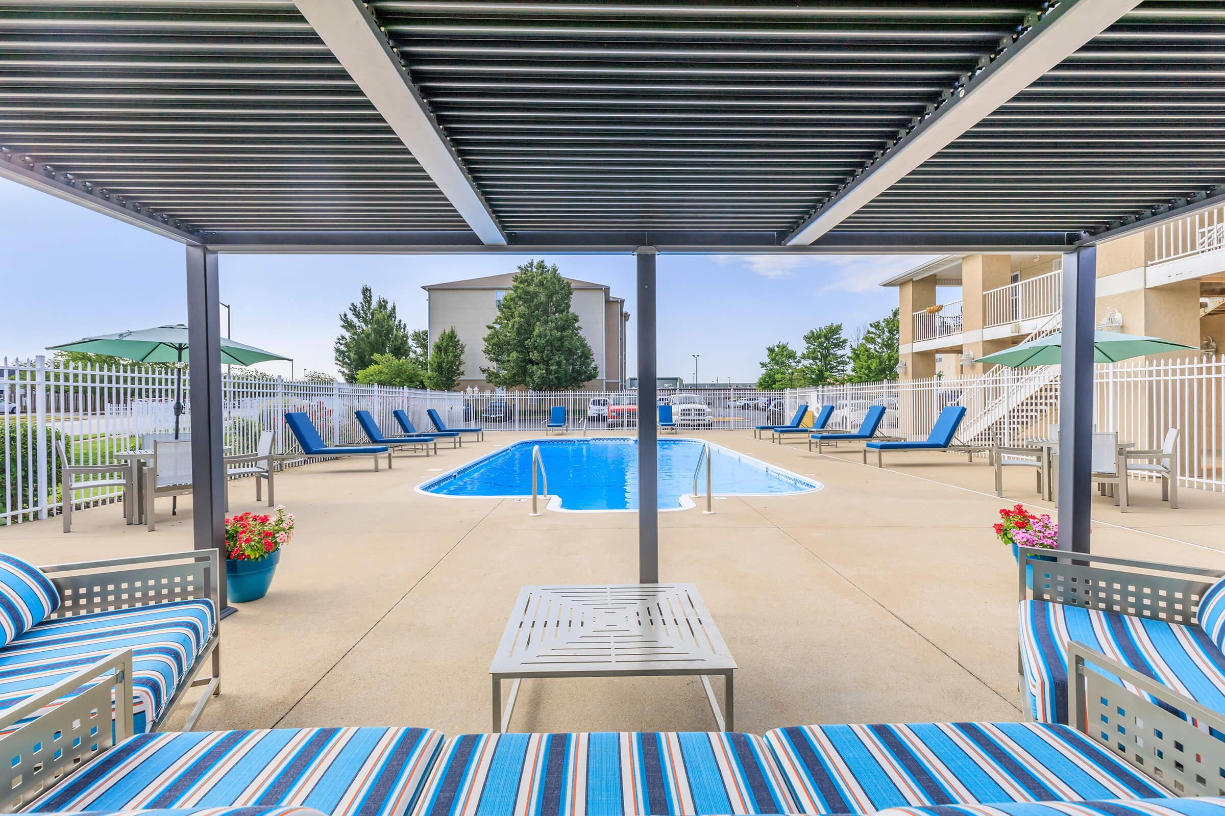 A view of a swimming pool surrounded by lounge chairs and shaded seating areas. The pool is set in a spacious outdoor area with potted flowers, umbrellas, and a fence in the background, offering a relaxing atmosphere for leisure and sunbathing.