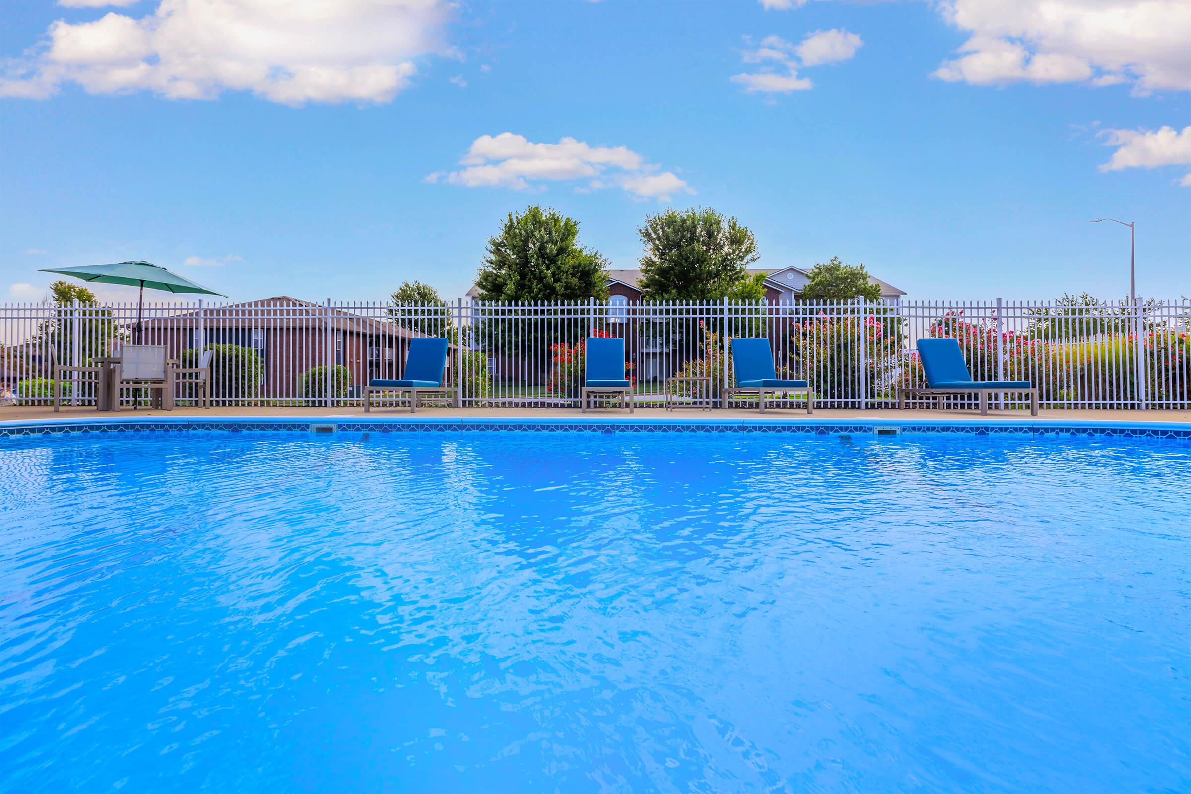 A clear blue swimming pool surrounded by a white fence, with several lounge chairs positioned nearby. In the background, there are green trees and residential buildings under a bright blue sky with some clouds. The scene conveys a relaxing outdoor atmosphere.