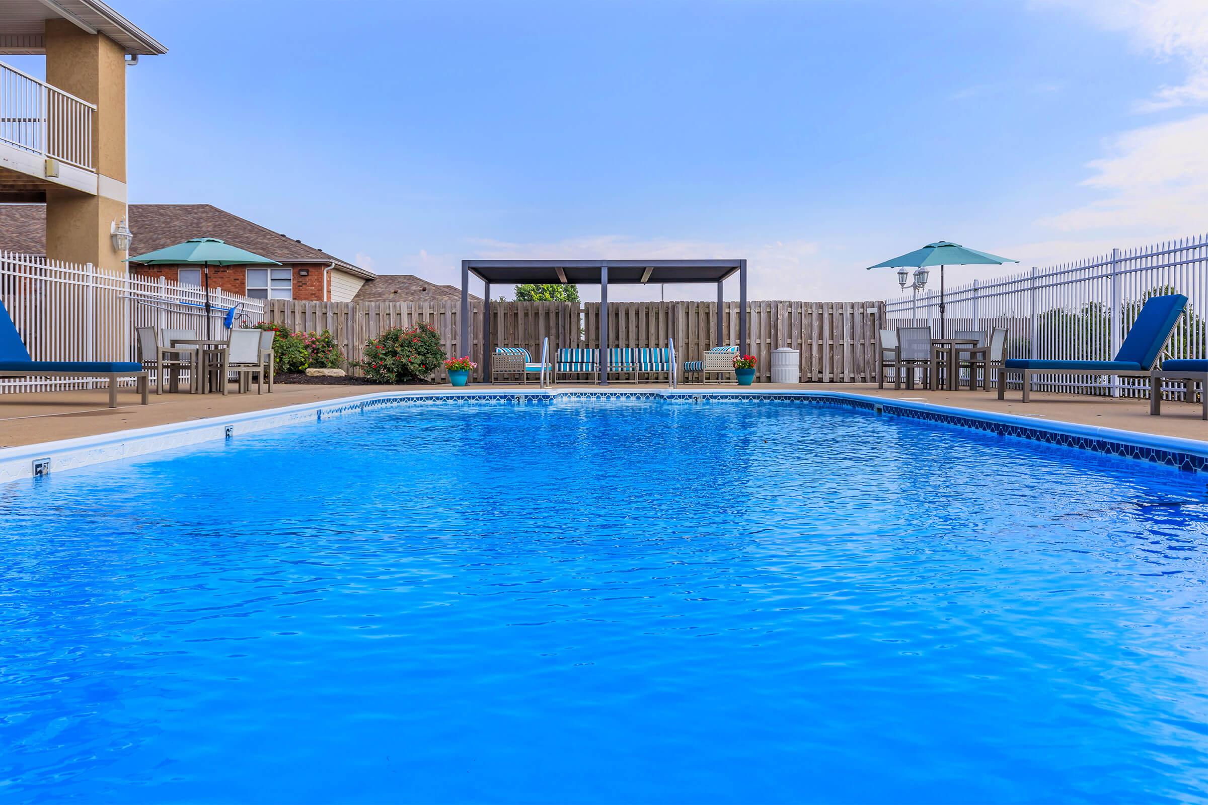 A clear blue swimming pool surrounded by a wooden deck. In the background, there are a few lounge chairs and tables, along with two green umbrellas. A shaded seating area is visible near the fence, and there are potted plants along the perimeter. The sky is mostly clear with a few clouds.