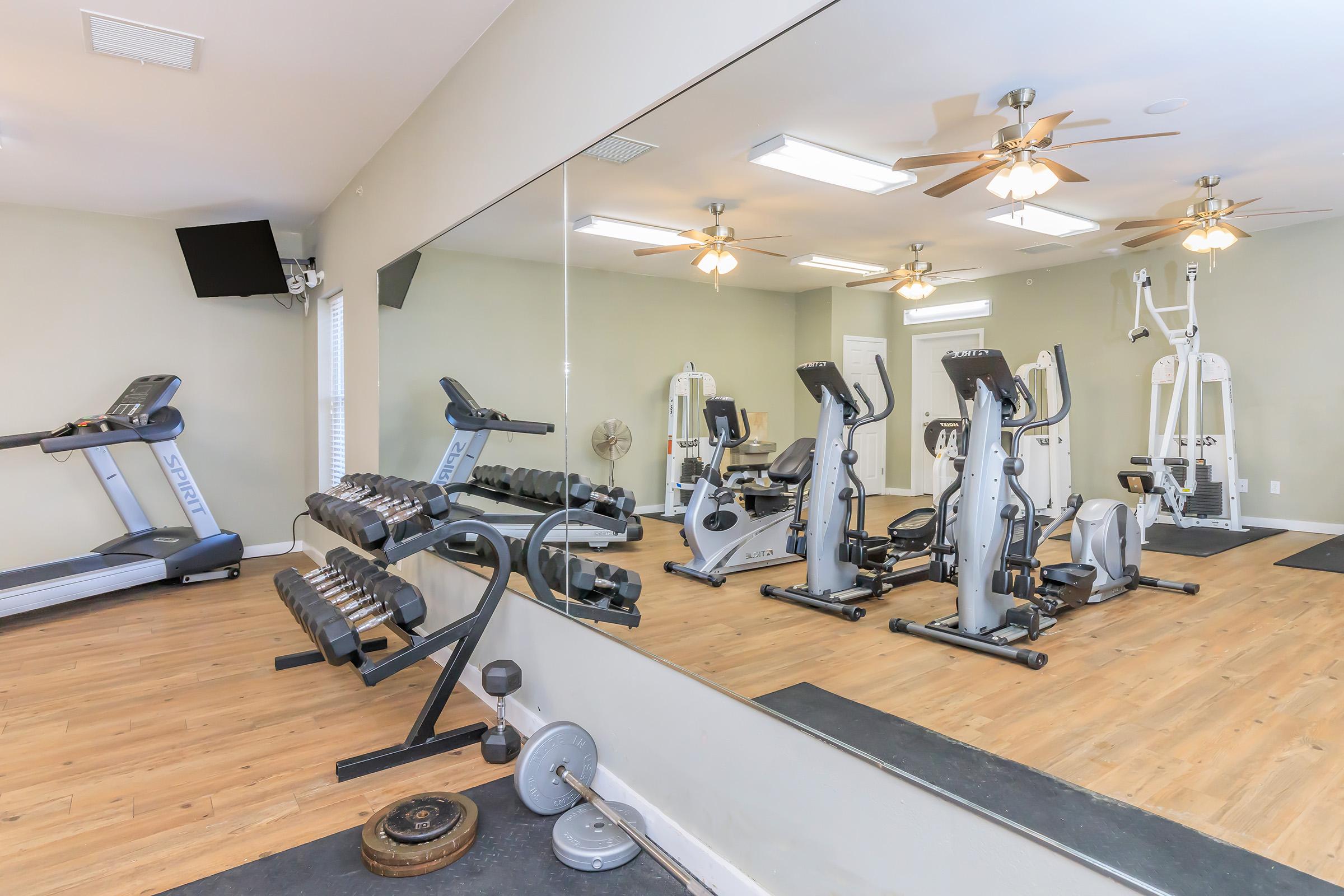 A modern gym interior featuring exercise equipment including treadmills, elliptical machines, weight benches, and a set of dumbbells. There are ceiling fans and mirrors on the walls, with a wooden floor providing a spacious and well-lit workout environment.