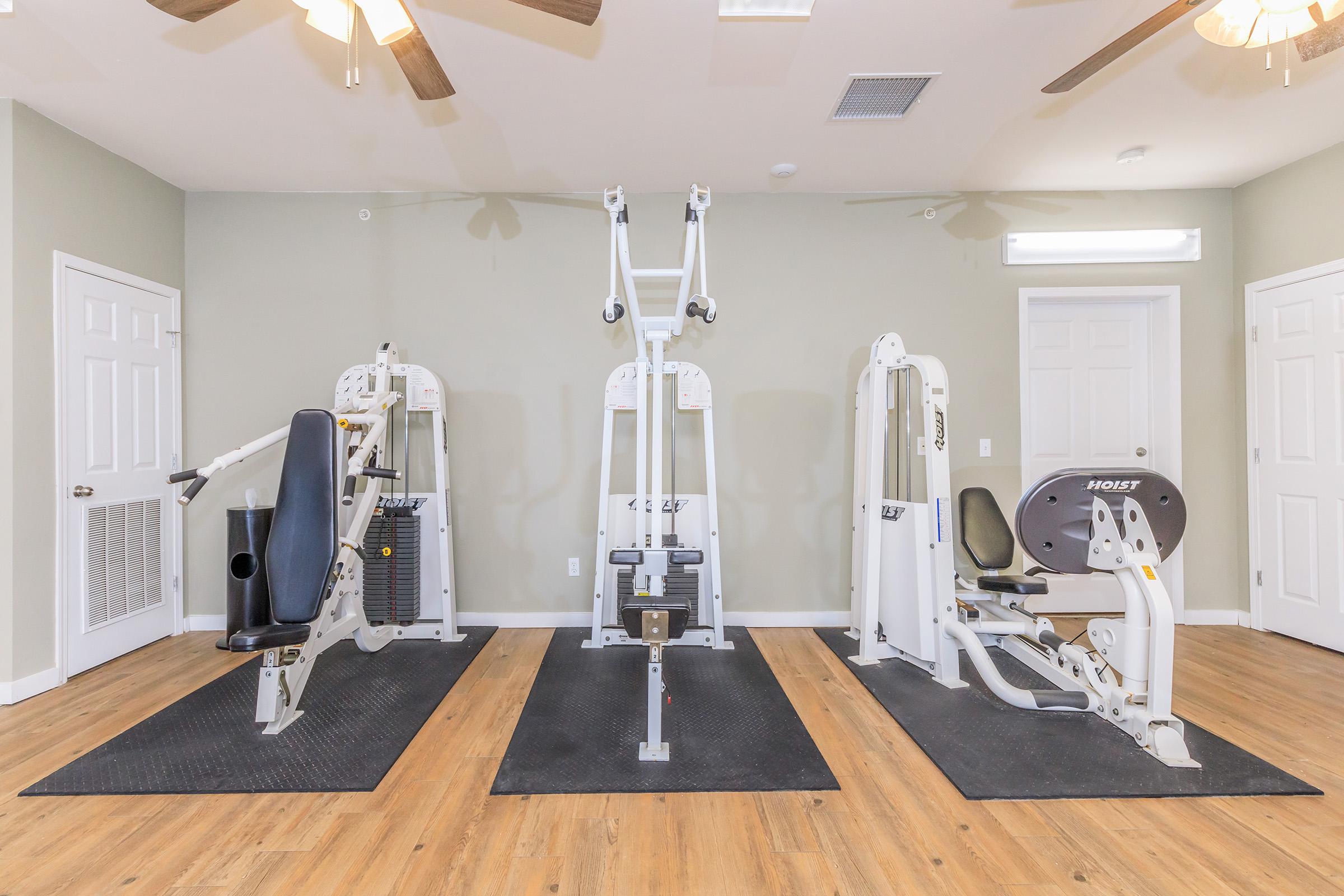 A well-equipped gym area featuring three exercise machines on a wooden floor, with a neutral-colored wall in the background. The machines include a seated leg press, a cable machine, and a seated chest press. Ceiling fans are visible, providing ventilation in the workout space.