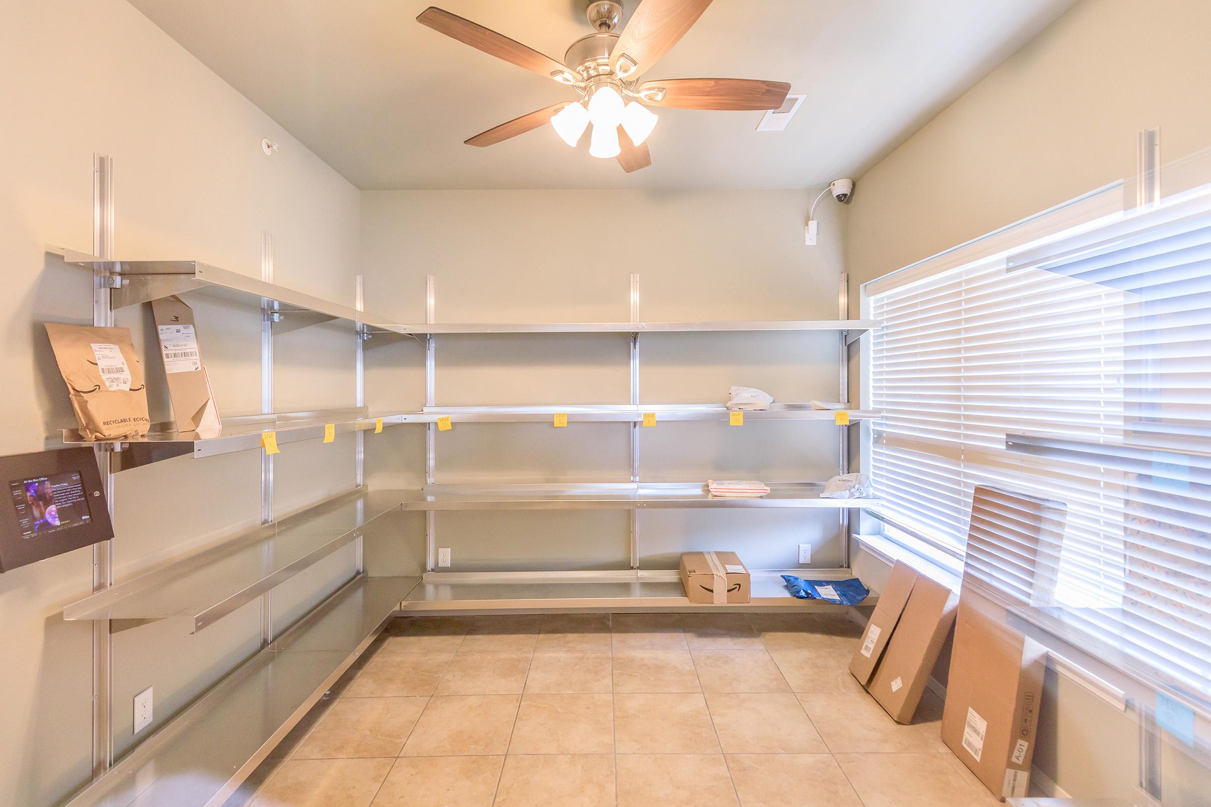 Empty room with gray walls and a ceiling fan. Shelving units along the walls are mostly bare, with a few items, including packages and paper, scattered sparsely. A bright window with blinds lets in natural light, and cardboard boxes are placed on the floor. The floor is tiled.
