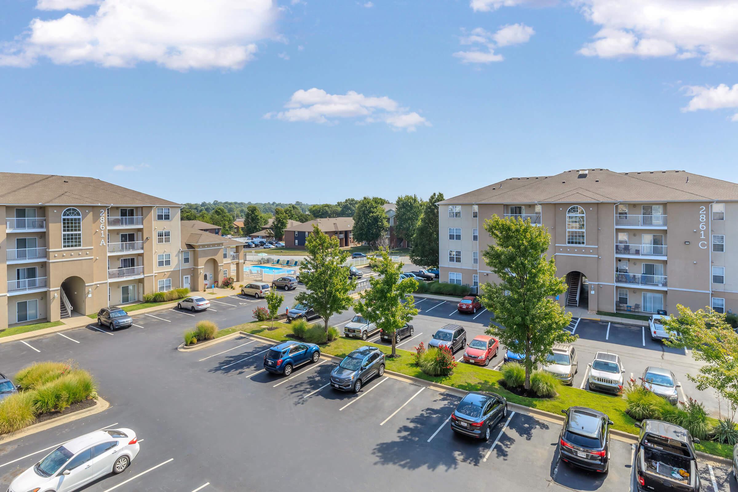 A view of a residential apartment complex featuring two multi-story buildings, surrounded by well-maintained landscaping and parking areas filled with cars. In the background, there is a blue sky with few clouds and a glimpse of a pool area.