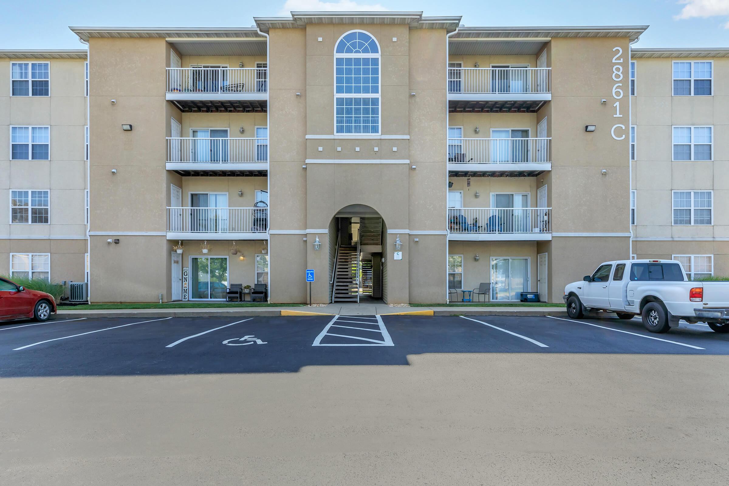 A three-story apartment building featuring a central archway entrance, balconies on the upper floors, and parked vehicles in the foreground. The building has a light brown exterior and is located in a paved parking lot with designated spaces, including a handicap-accessible spot.