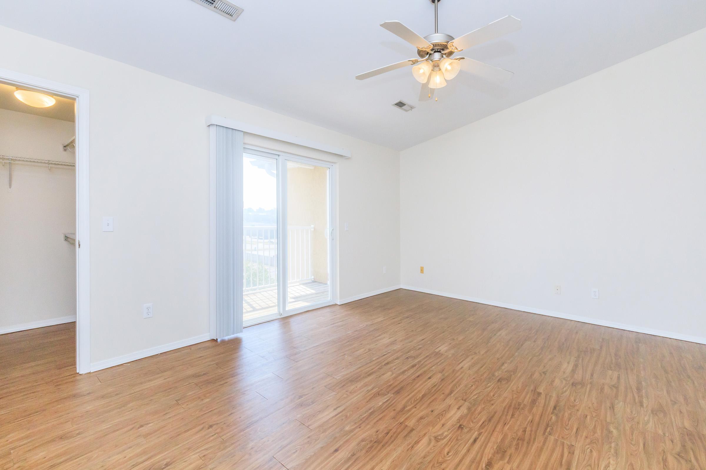 Empty room with light wood flooring and a ceiling fan. There is a sliding glass door leading to a balcony, and a small closet is visible on the left side. The walls are painted in a light color, giving the room a bright and spacious feel.