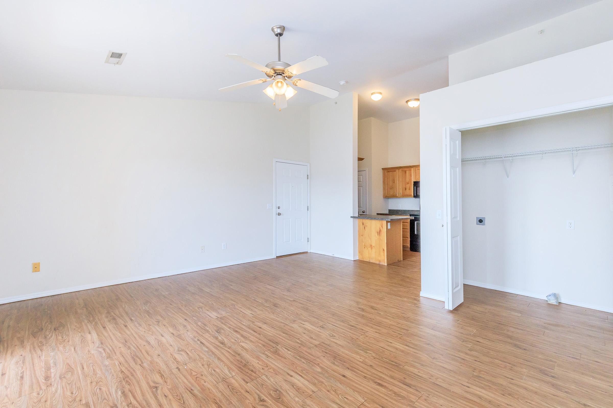 A spacious, bright living area with light-colored walls and wood flooring. A ceiling fan hangs in the center, and a doorway leads to a kitchen area with wooden cabinets visible in the background. An empty closet is situated to the right, creating an open and inviting ambiance.