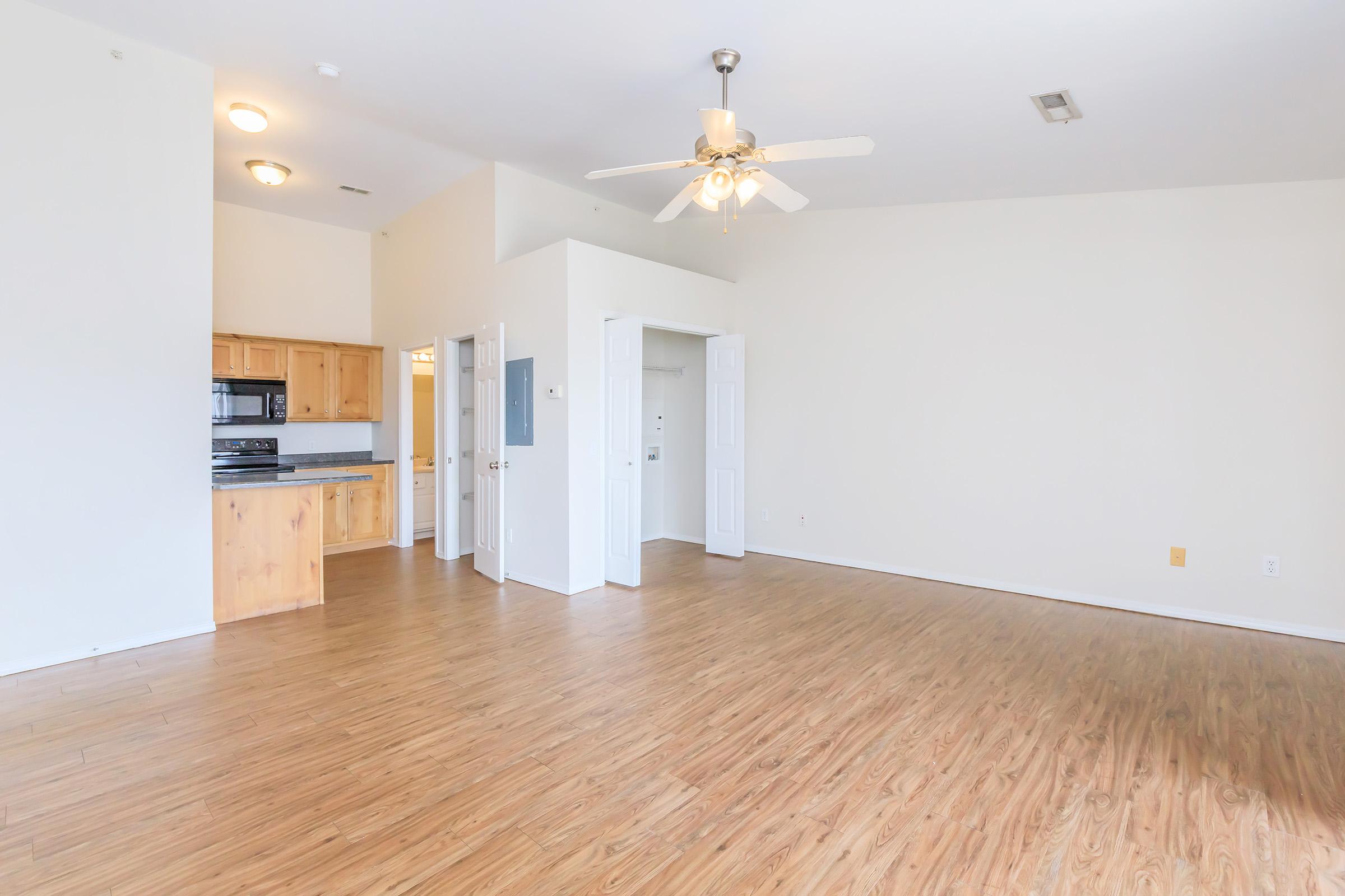 Spacious living area featuring light-colored walls and hardwood flooring, with a ceiling fan and natural light. The kitchen area is partially visible, equipped with wooden cabinets and modern appliances. Two doorways lead to other rooms, creating an open and airy layout.