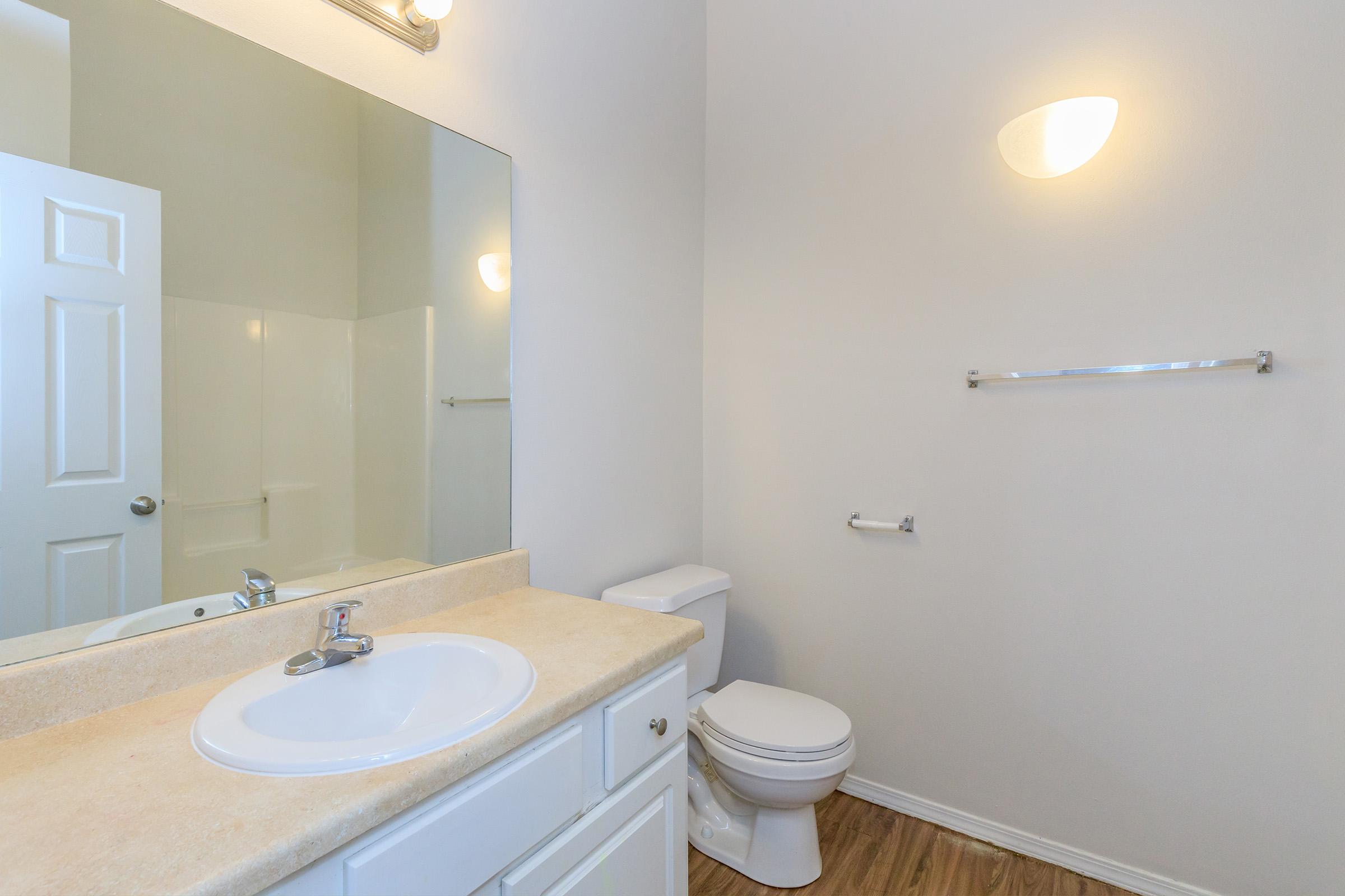 A clean and modern bathroom featuring a countertop sink with a faucet, a large mirror above the sink, a toilet, and a shower area. The walls are painted in light colors, and there is a towel rack on one side. The flooring is a warm wood tone, creating a bright and inviting space.