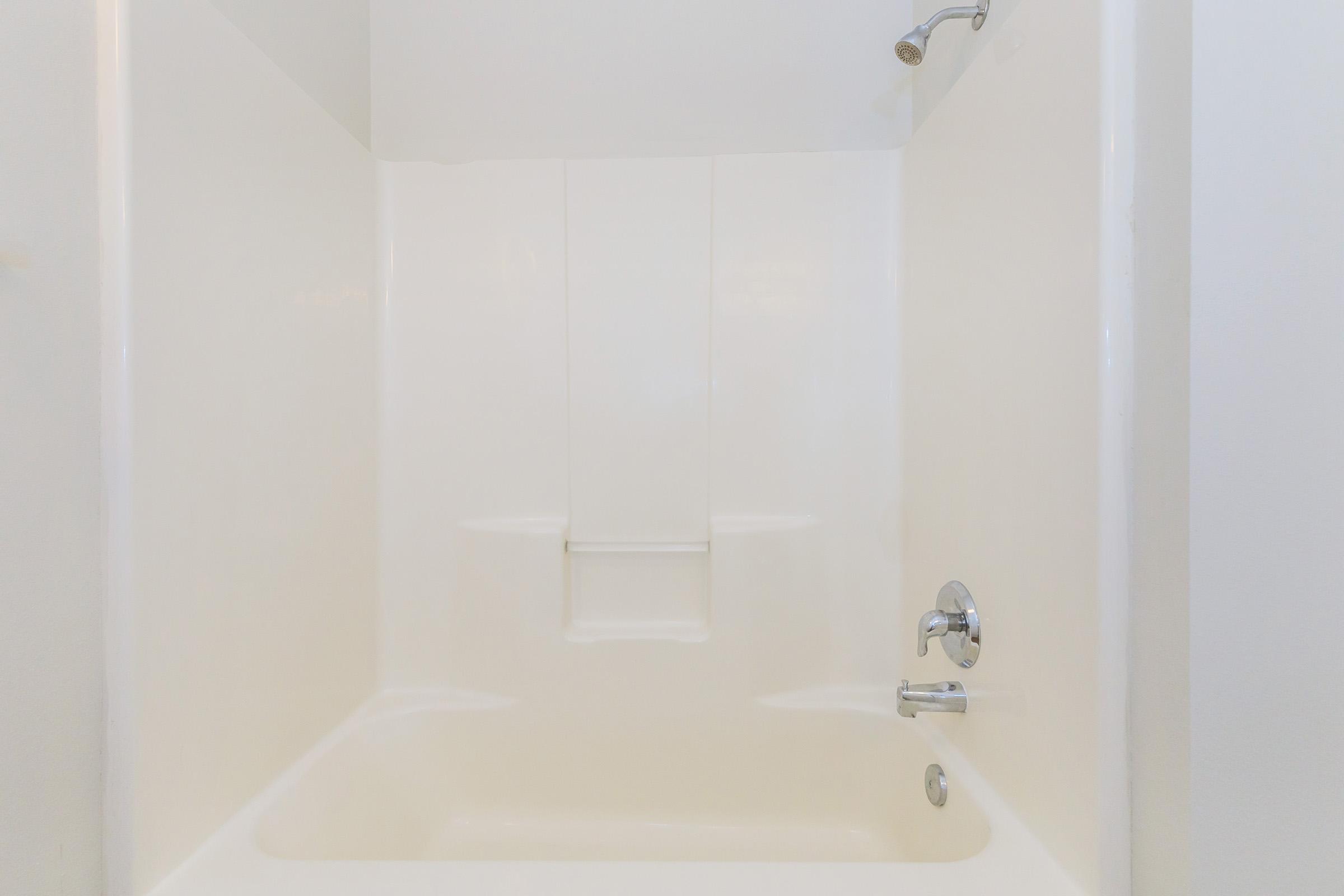 A clean, empty bathtub with white walls and a simple design. The faucet and showerhead are visible, along with a minimalistic grab bar on the wall. The space is well-lit, indicating a fresh and airy bathroom environment.