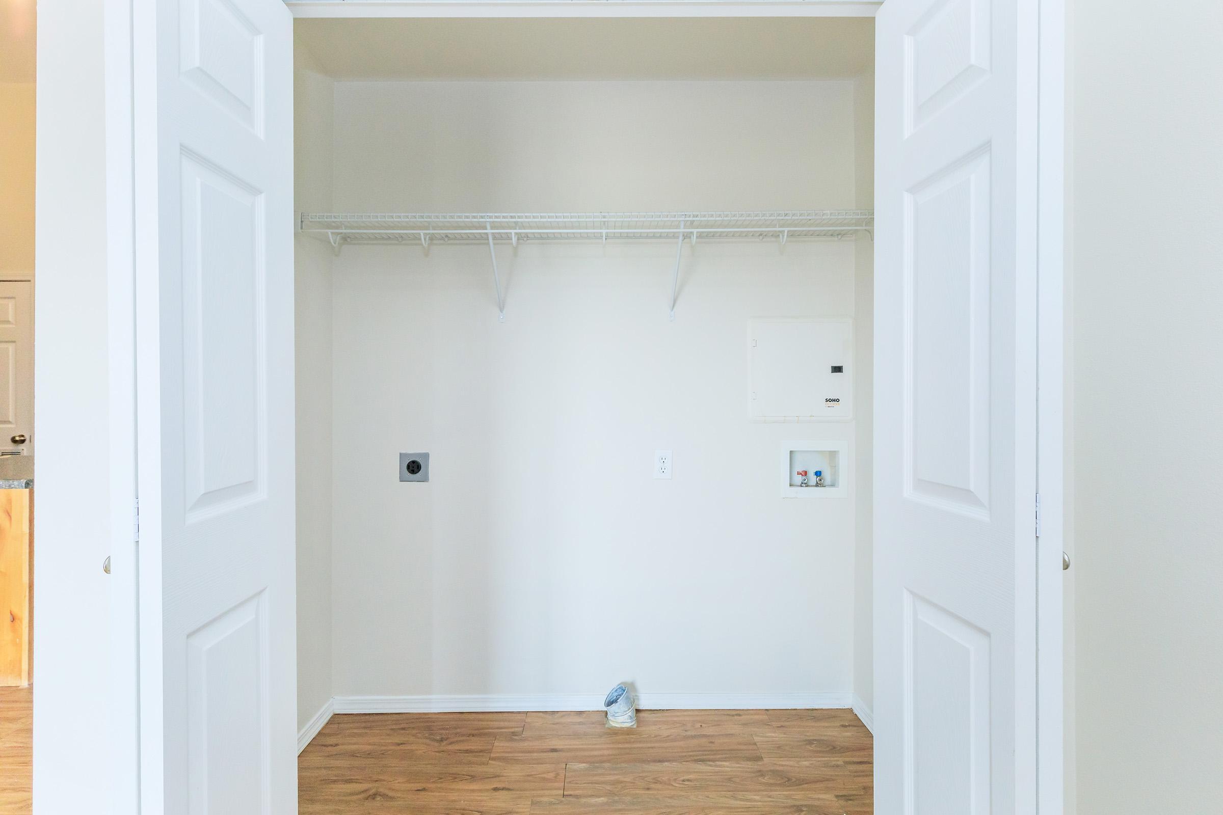 An empty closet with two white doors open, revealing a light beige wall and a wooden floor. A wire shelf runs along the back wall, and the only items visible are a small vent and an electrical panel on the left side. The overall space is bright and uncluttered.