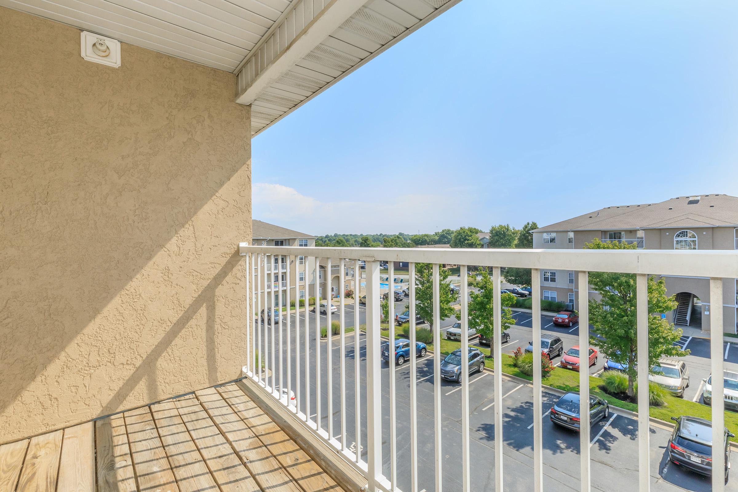 View from a balcony overlooking a parking lot and nearby apartment buildings. The sky is bright and clear, with a few scattered clouds. Green trees line the area, providing a pleasant atmosphere. The balcony has wooden flooring and a white railing.