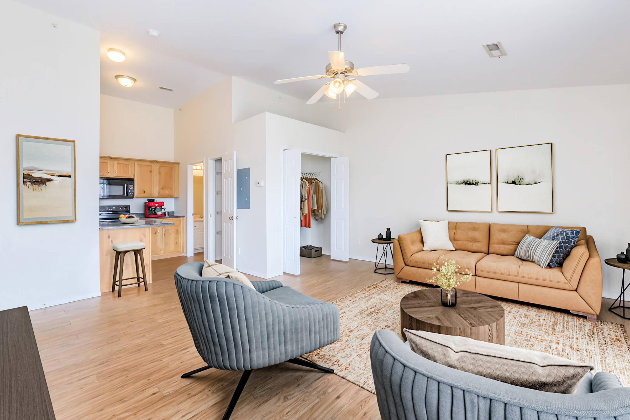 A modern living room featuring a tan sofa, two gray armchairs, and a round wooden coffee table. The walls are painted white with two framed artworks. A kitchen area is visible in the background, and a closet with casual clothes is slightly open. Natural light fills the space, accentuating the warm, inviting atmosphere.