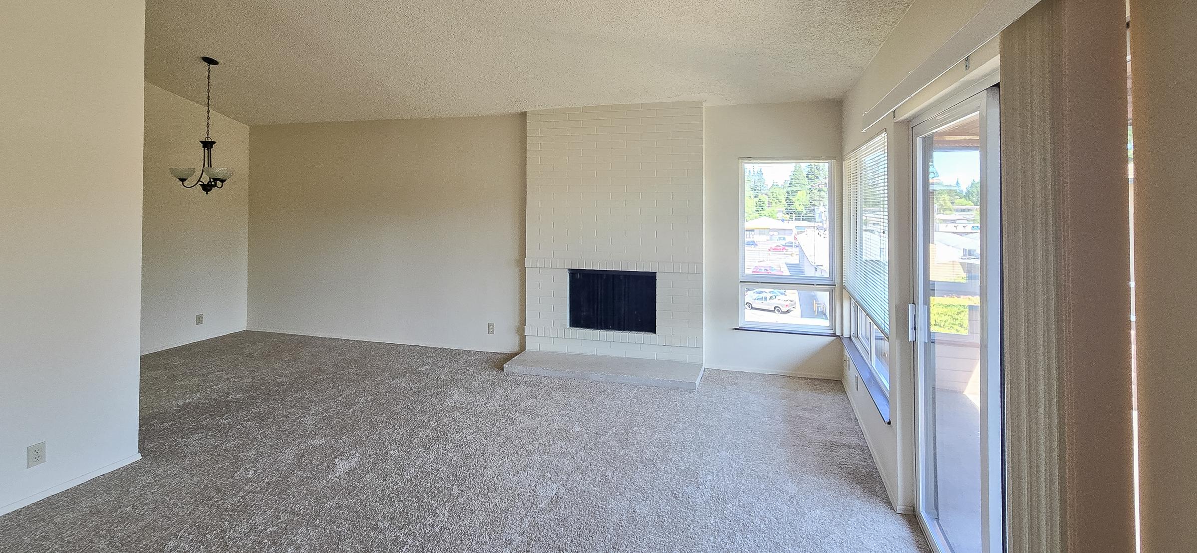 Spacious living room featuring a beige carpet, a white-brick fireplace, and large windows with natural light. A pendant light hangs from the ceiling, and there's a sliding glass door leading to an outdoor area. The walls are painted in a light color, creating a bright and airy atmosphere.