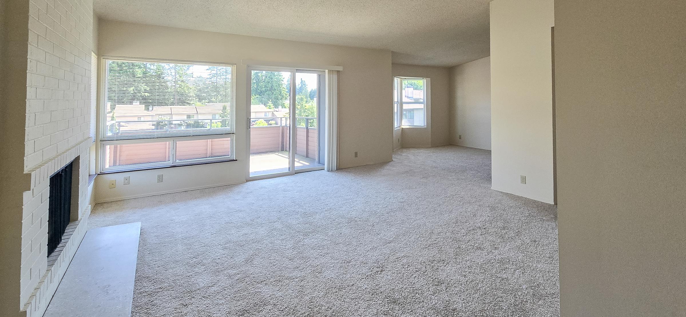 Spacious and bright living room with large windows allowing natural light to fill the space. Features a beige carpet, a white brick fireplace on one wall, and a sliding glass door leading to a balcony. The walls are painted in a light color, enhancing the airy feel of the room.