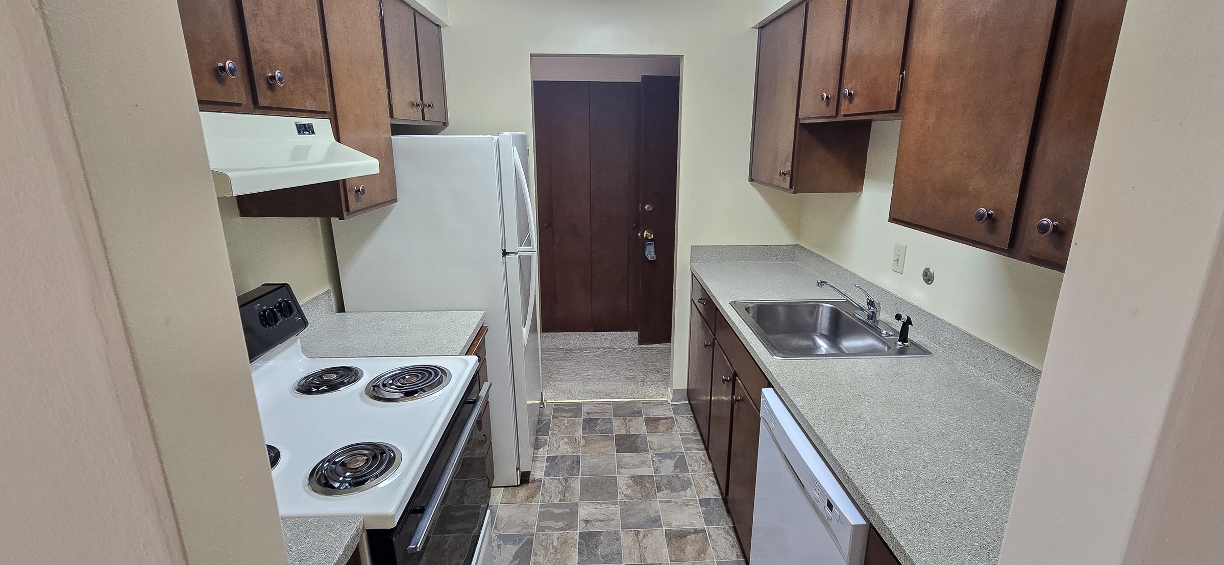 A small, functional kitchen featuring wooden cabinets, a countertop with a sink, an electric stove, a refrigerator, and a door leading to another room. The floor is tiled, and there is ample natural light illuminating the space.