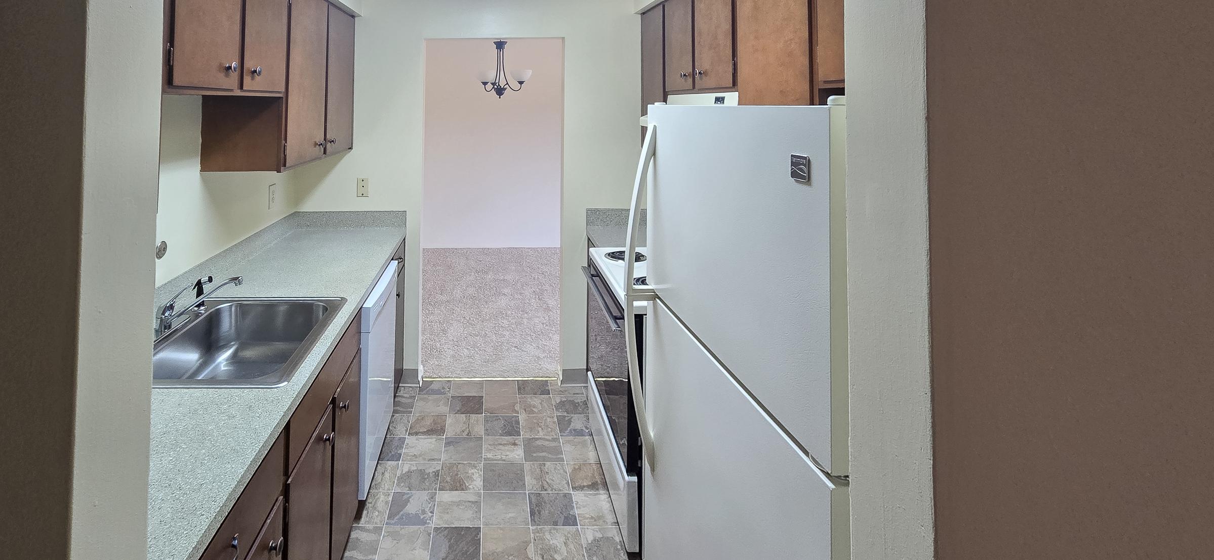 A view of a kitchen showing wooden cabinets, a double sink, a dishwasher, and a white refrigerator. The flooring features tiled squares, and there is a doorway leading to another room. A chandelier hangs from the ceiling, providing light to the space.