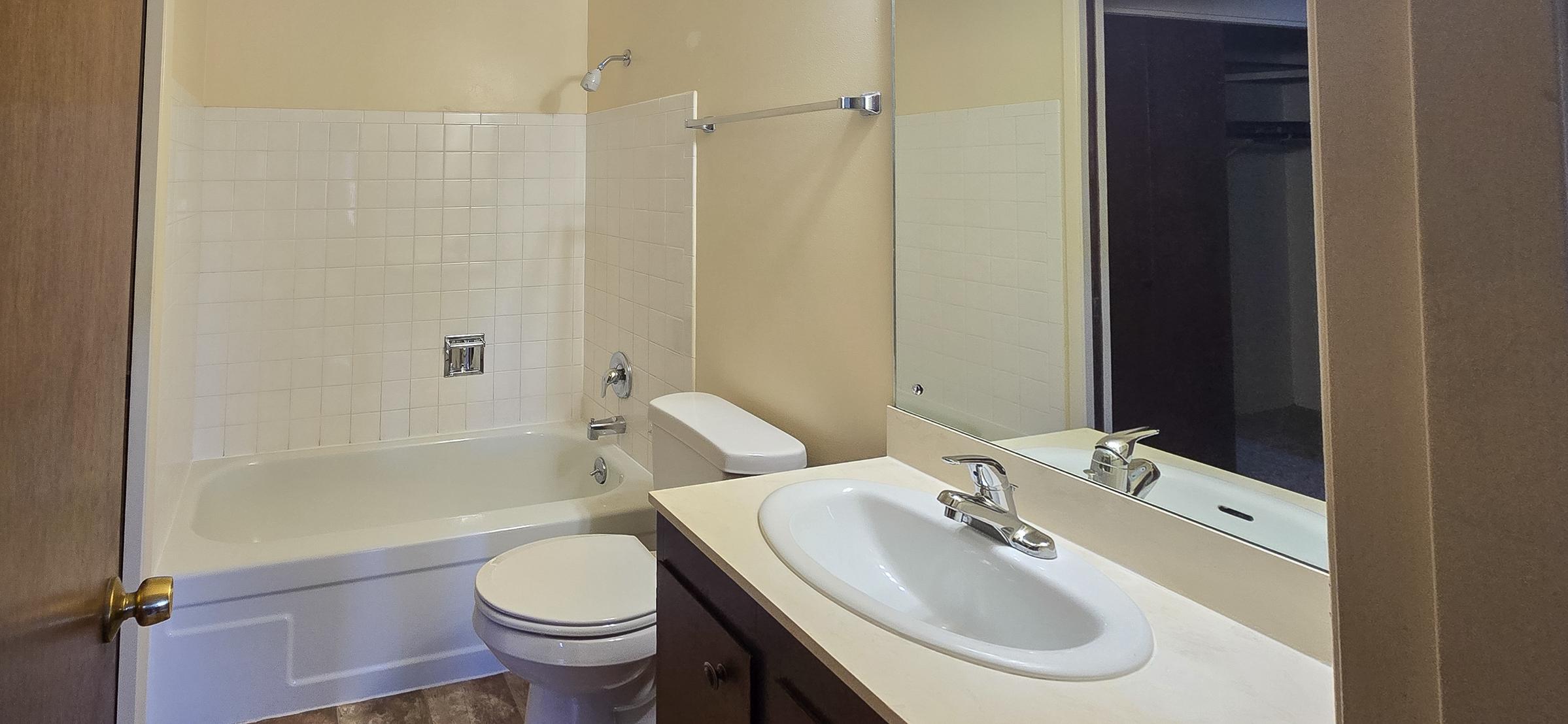 A clean, well-lit bathroom featuring a bathtub with a shower, a toilet, and a sink with a mirror above it. The walls are pale, and there are white tiles around the bathtub area. A wooden door leads to another room, with a closet visible in the background.
