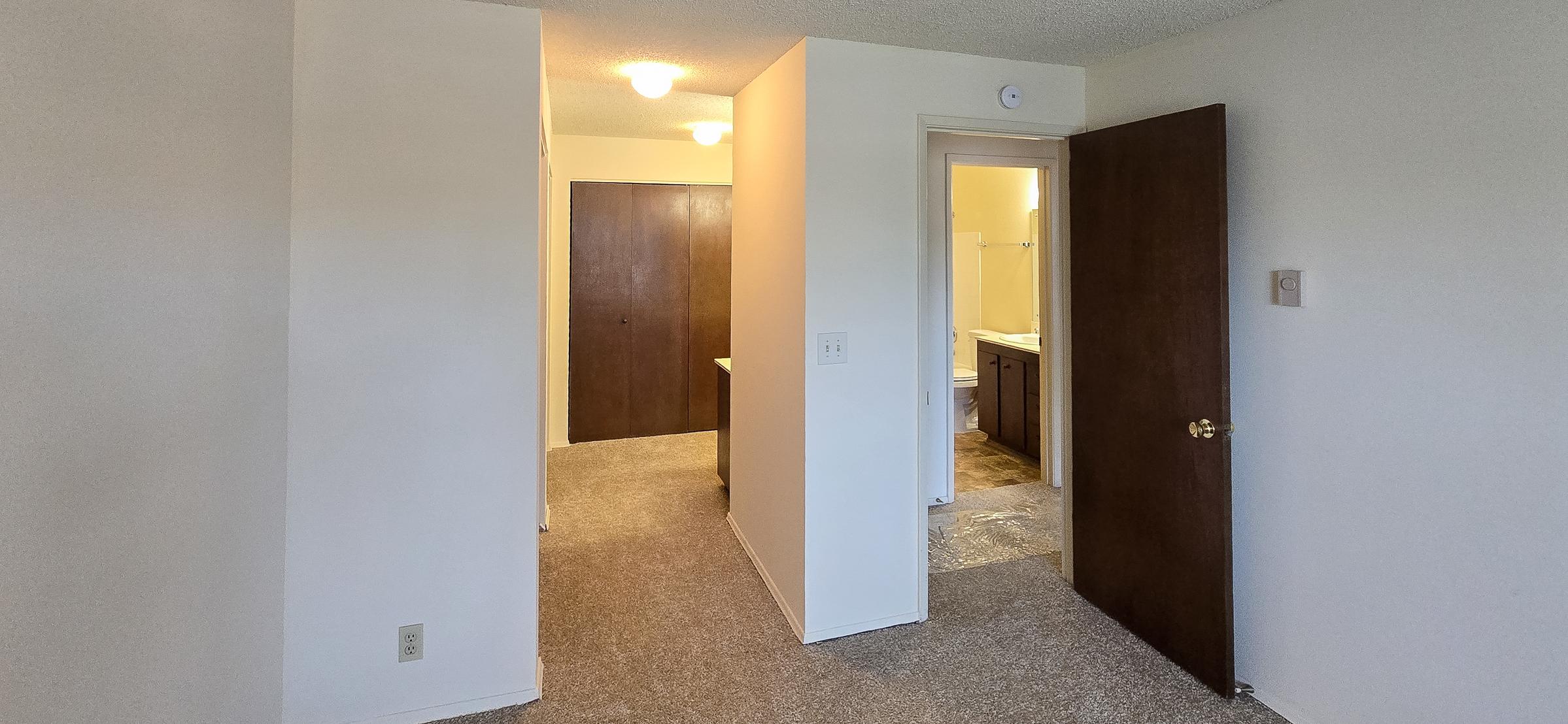 Interior view of a residential space showing a hallway with beige walls and carpet. On the left, there's a closed wooden door, and at the end of the hallway, an open door leads to a bathroom. Light fixtures are visible on the ceiling, providing illumination. The space appears clean and unoccupied.