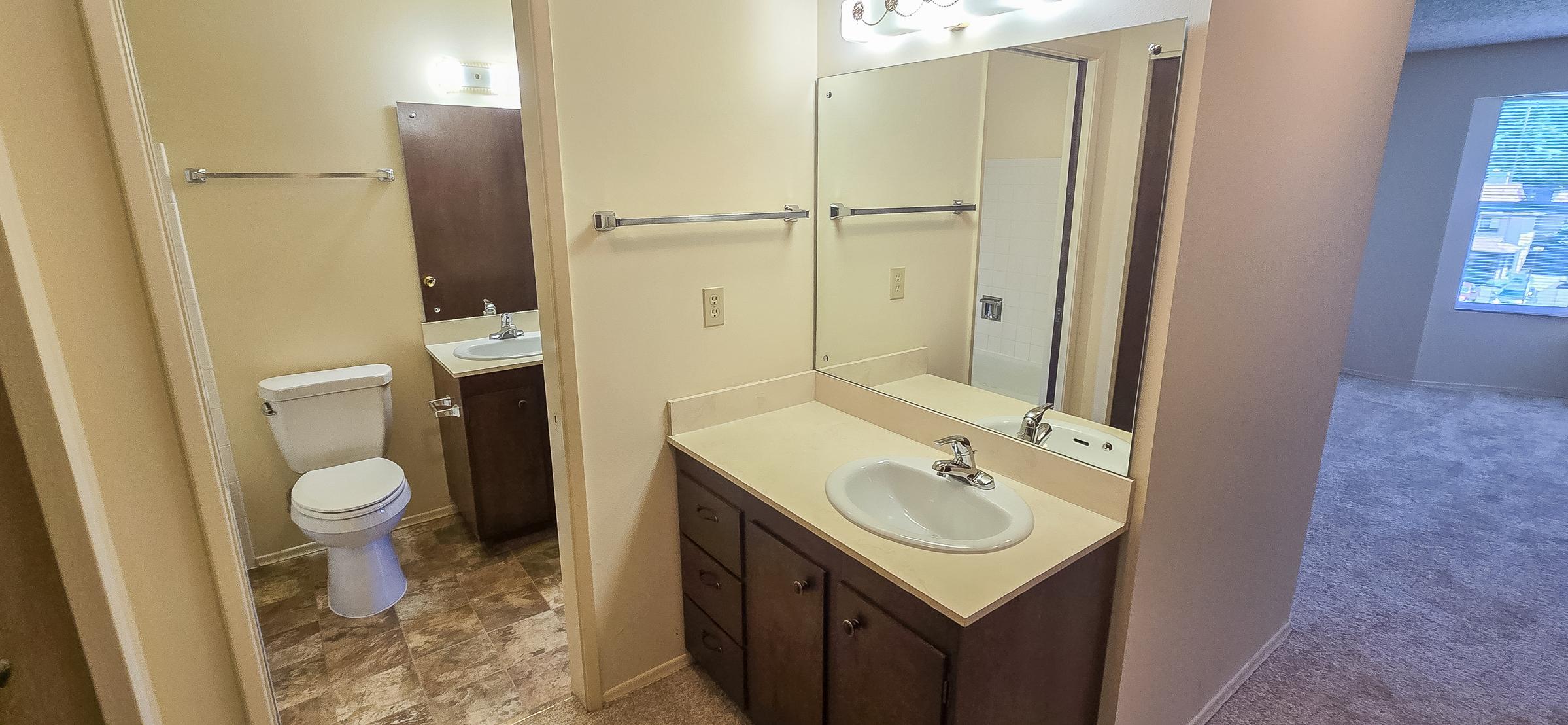 A bathroom featuring a beige countertop with a sink, a large mirror, and wooden cabinetry. To the left, there is a toilet and a wall-mounted towel rack. The flooring is tile, and the walls are painted a light color. In the background, there is a glimpse of a carpeted room with a window.