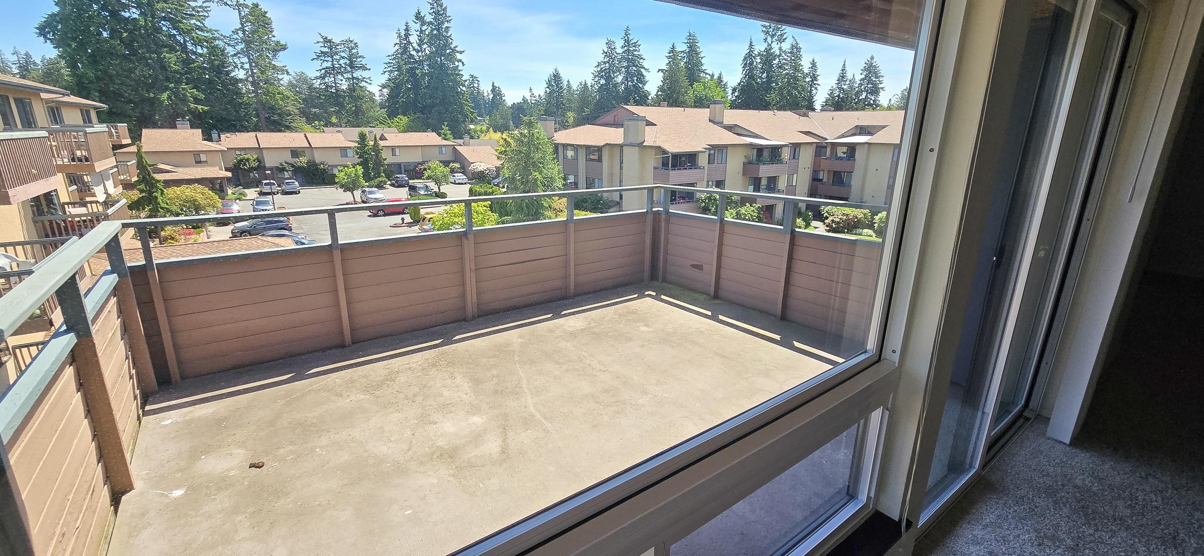 View from a balcony overlooking a well-maintained residential complex. The scene features a clear blue sky, greenery from surrounding trees, and an asphalt parking area filled with cars. The balcony has a concrete floor and a railing, providing an open and airy atmosphere.