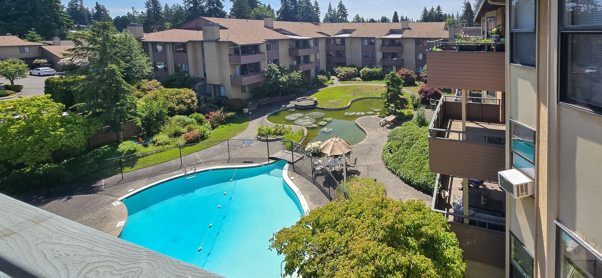 Aerial view of a residential complex featuring a large swimming pool and landscaped gardens with walking paths, benches, and water features. Surrounding buildings are three stories high, set among lush greenery and trees, creating a serene living environment. Clear blue sky in the background.