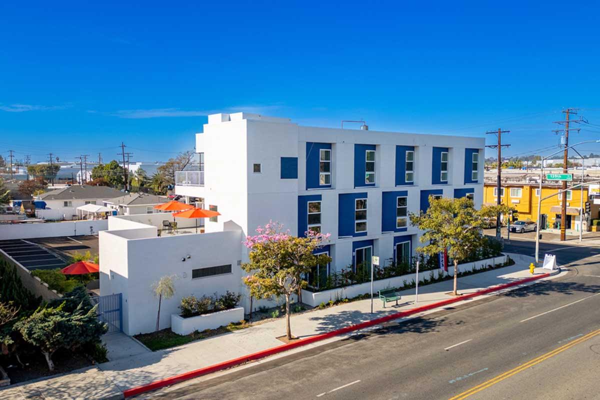 A modern three-story building with blue and white exterior, featuring large windows and a landscaped front yard. The street is lined with trees and there are orange umbrellas in an outdoor seating area. Clear blue sky in the background, with power lines visible.