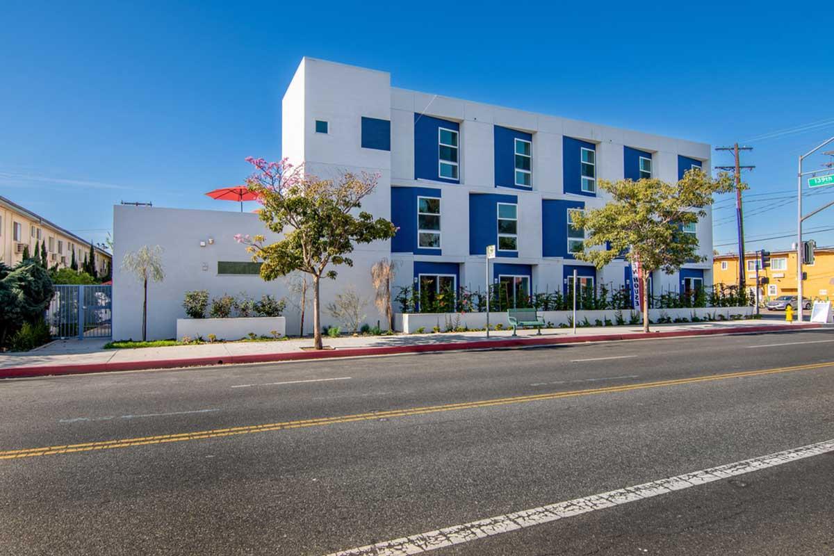 A modern, three-story building featuring a white facade with blue accents. The structure has several windows, some greenery in front, and a red umbrella visible on the rooftop. The scene is set on a sunny day, with a wide street in the foreground and clear blue skies above.