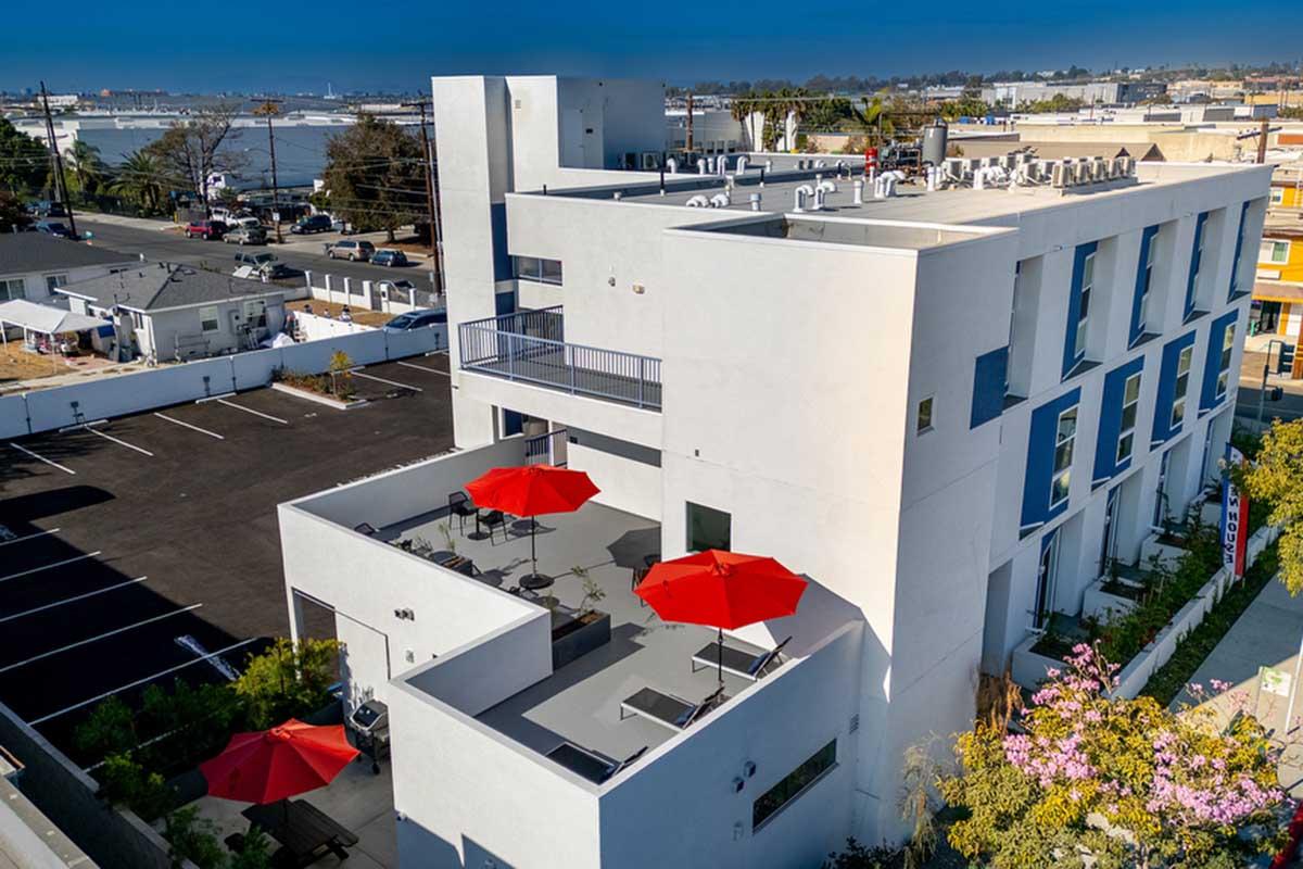 Aerial view of a modern building featuring a rooftop terrace with red umbrellas. Surrounding area includes parked cars and other buildings, with a clear sky in the background. The design is contemporary with clean lines and a light color palette.