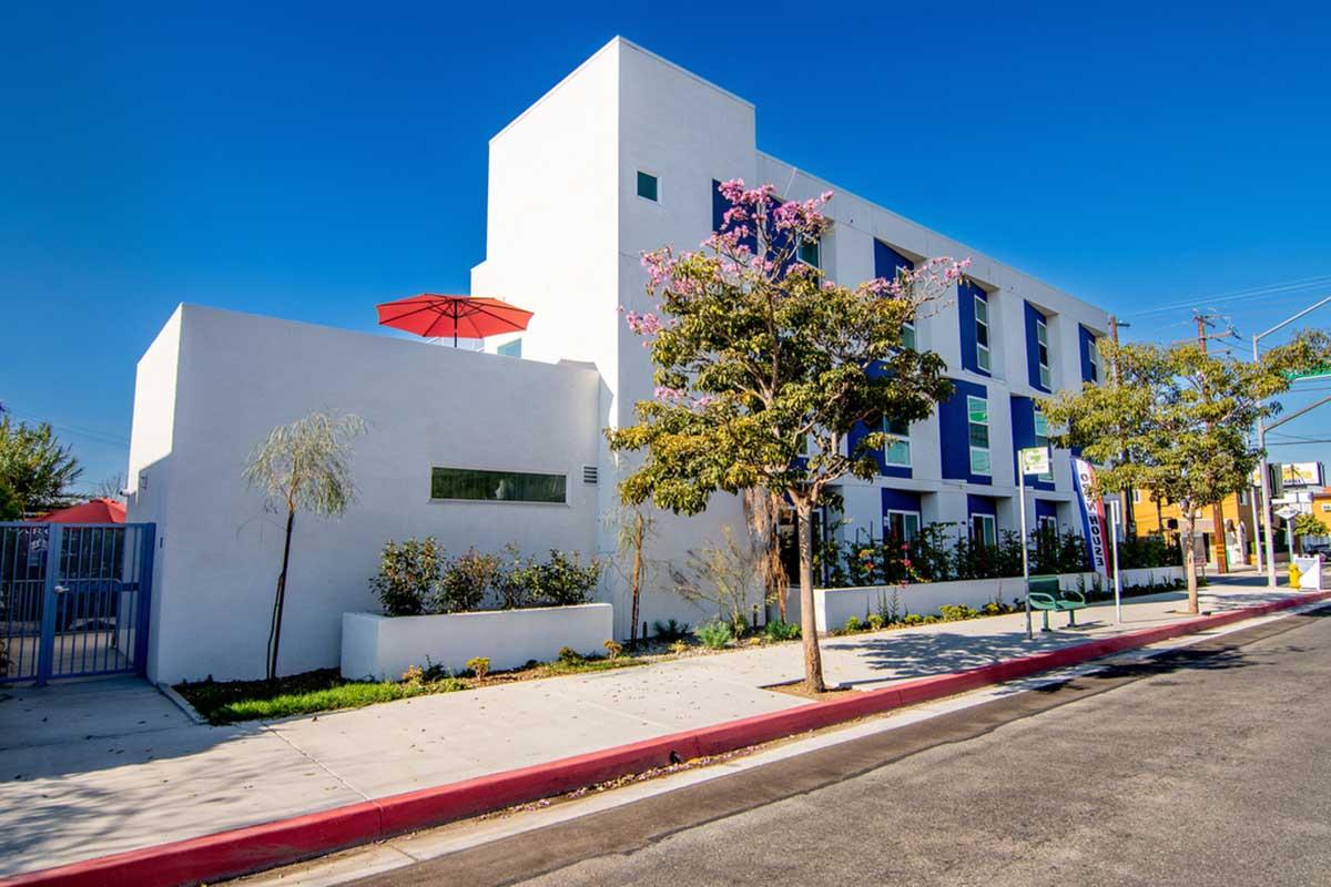 Modern building with a white exterior and colorful accents, featuring a rooftop terrace with a red umbrella. The area includes landscaped greenery and a sidewalk along a quiet street under a clear blue sky.