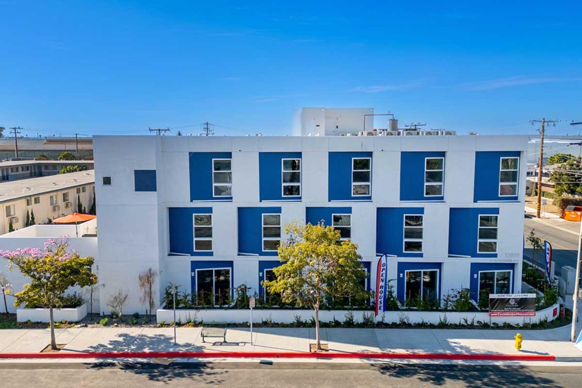 A modern three-story building with a white exterior and dark blue accents. Large windows are lined in rows across the front. Decorative landscaping is visible in the foreground, with a sidewalk and street in front. Clear blue sky above enhances the bright appearance of the building.