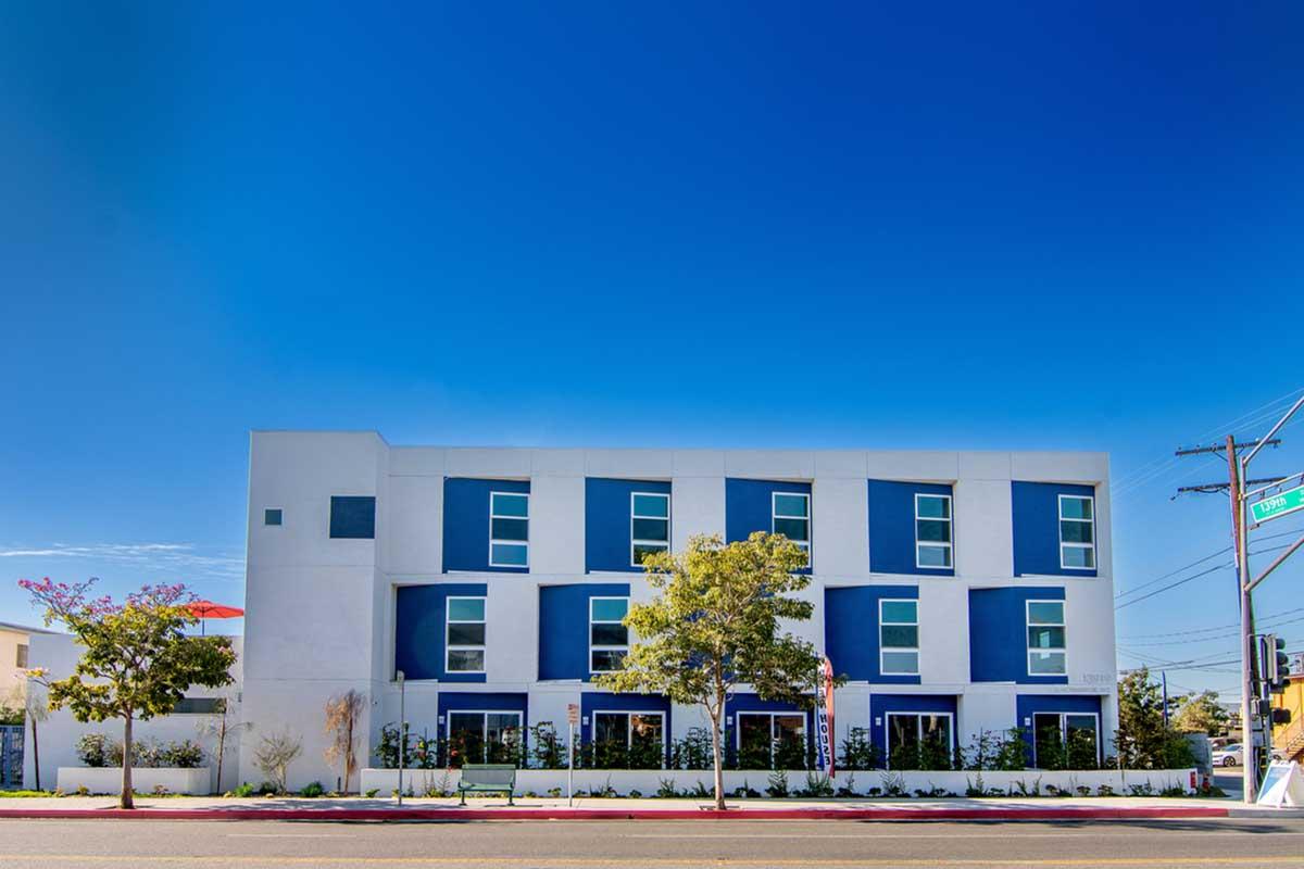 Modern three-story building with a white and blue facade, featuring large windows and landscaped greenery at the base. The sky above is clear and blue, with a few electrical poles visible on the side. The scene depicts a well-maintained urban environment.