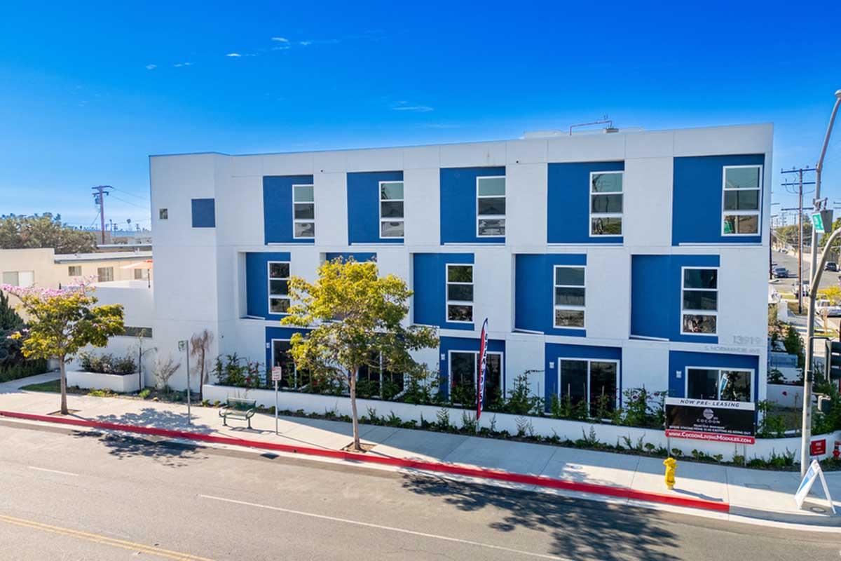 Modern multi-story building with a predominantly white exterior and blue accents. Features large windows, a landscaped area in front, and trees. A flag is displayed outside, along with a banner on the façade. The scene is set on a clear day with a bright blue sky.