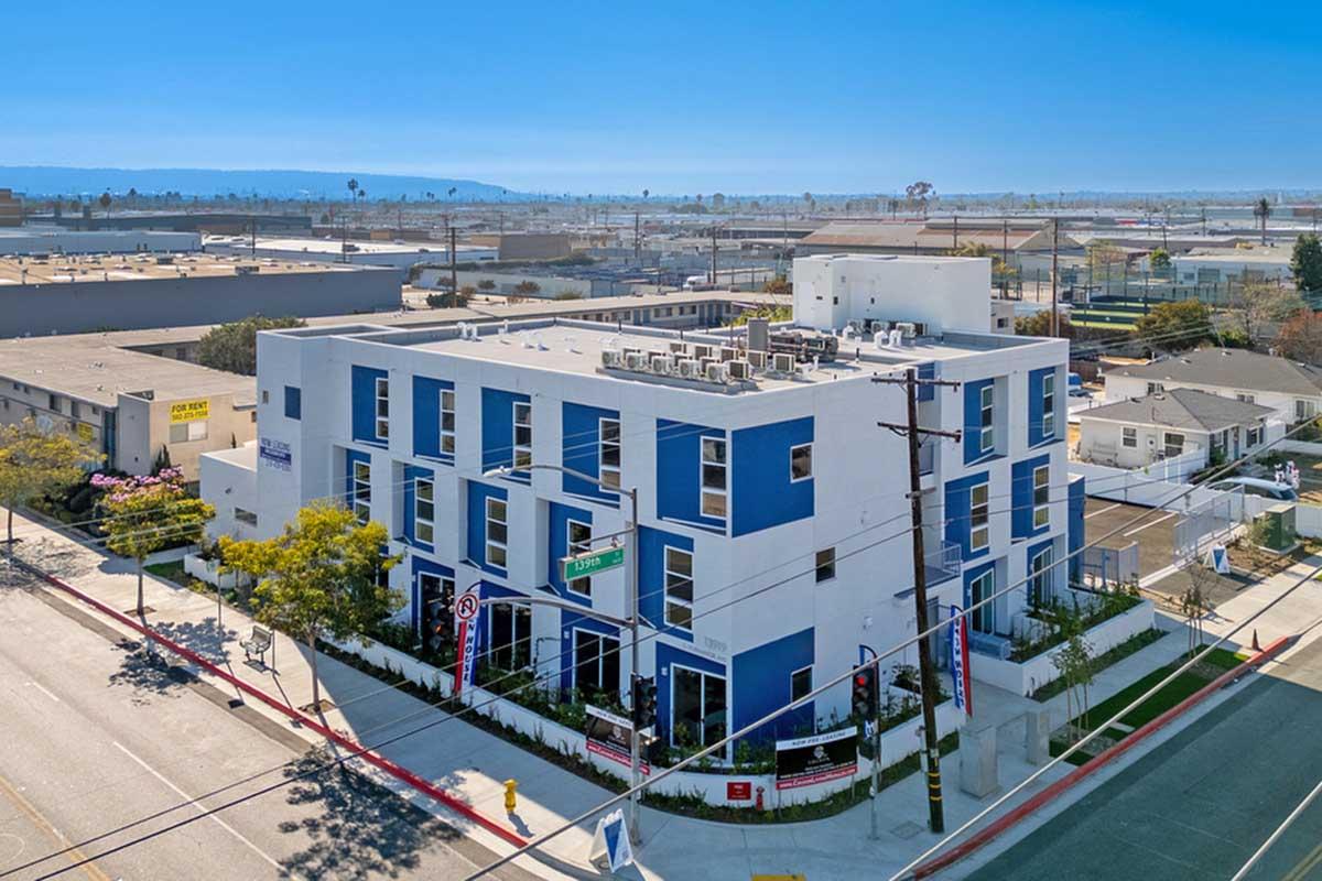 Modern multi-story building with a white and blue facade, featuring large windows and balconies. The surrounding area includes other commercial buildings and residential homes. Clear blue sky overhead, with power lines in the foreground and a landscaped section in front of the building.