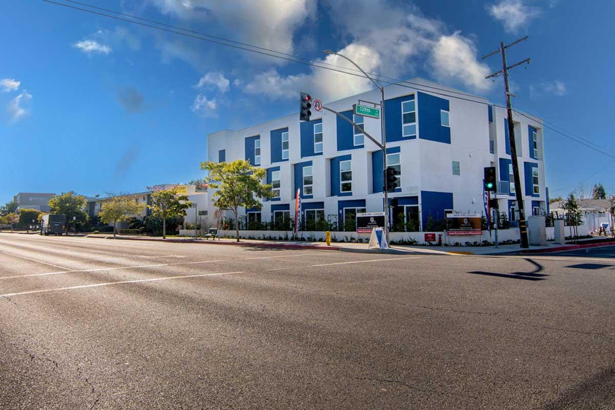 A modern building with a blue and white exterior is situated at a street corner. Traffic lights and road signs are visible, along with greenery and sidewalks. The scene is set under a clear blue sky with scattered clouds.