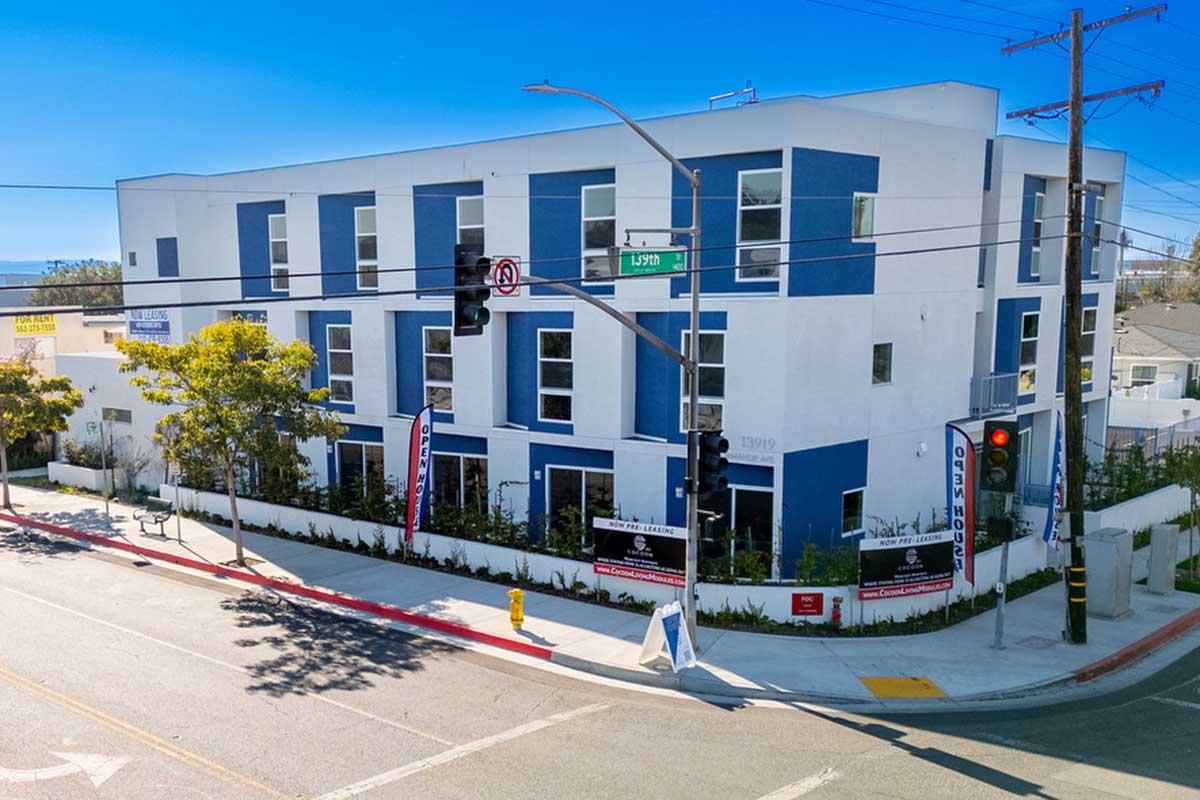 A modern, three-story building with a white and blue exterior design. It features large windows and multiple entrances. Surrounding the building are trees and signage, indicating it may be a commercial or residential space. Traffic lights and a sidewalk are visible in front of the property.