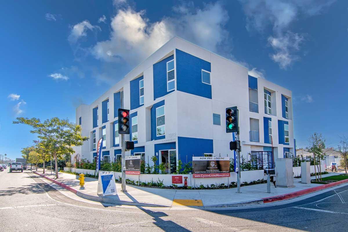Modern multi-story building with a blue and white facade, located at a street corner. Pedestrian traffic lights in the foreground and landscaped areas with trees and signage around the property. Clear blue sky with a few clouds.