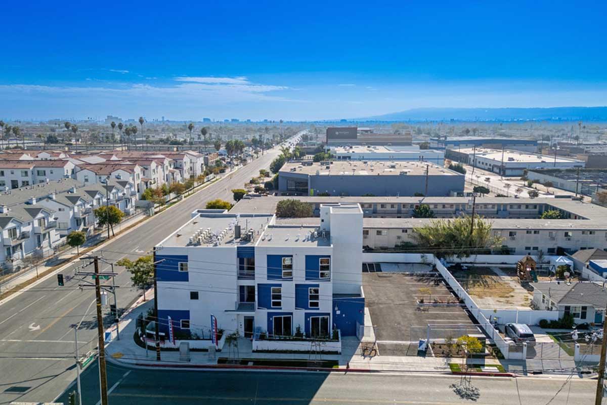 Aerial view of a suburban area featuring a mix of residential buildings and commercial properties. In the foreground, a modern white and blue building with a parking lot, surrounded by a mix of houses, streets, and distant mountains under a clear blue sky. Power lines run along the road.