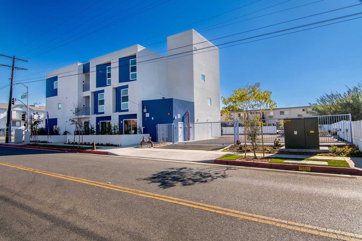 A modern, two-story building with a white and blue façade, surrounded by a white fence and landscaping. The street is visible in front, with utilities on the side and a clear blue sky overhead. The area appears well-maintained and inviting.
