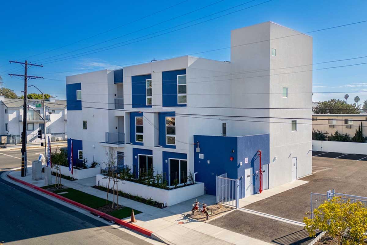 Modern multi-unit residential building featuring a mix of white and blue exterior, large windows, and a clean design. The building is located on a street with power lines, surrounded by a landscaped area and an adjacent parking lot. Clear blue sky is visible overhead.